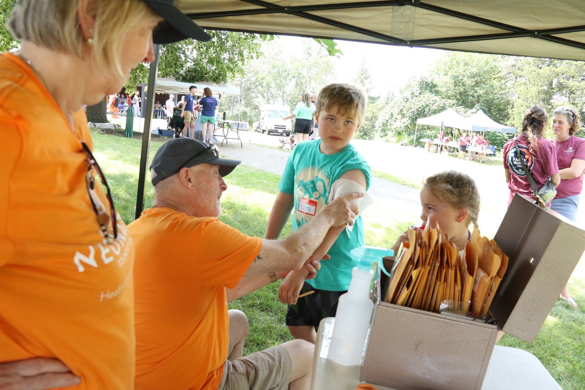 Man applying temporary tattoo to a child's arm at an outdoor event, other people in the background.