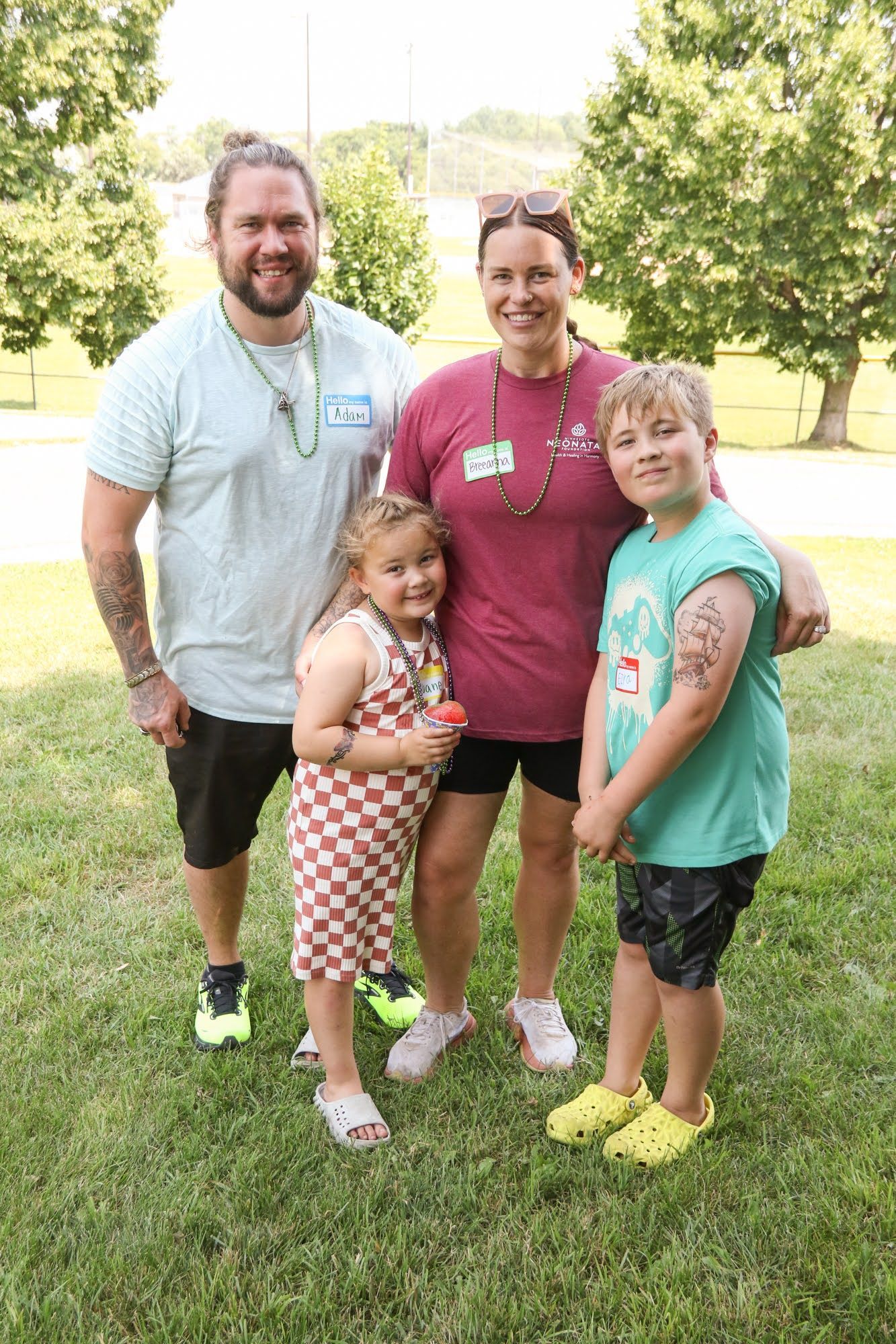 Family of four poses on grass: dad, mom, and two kids. Green trees in background.