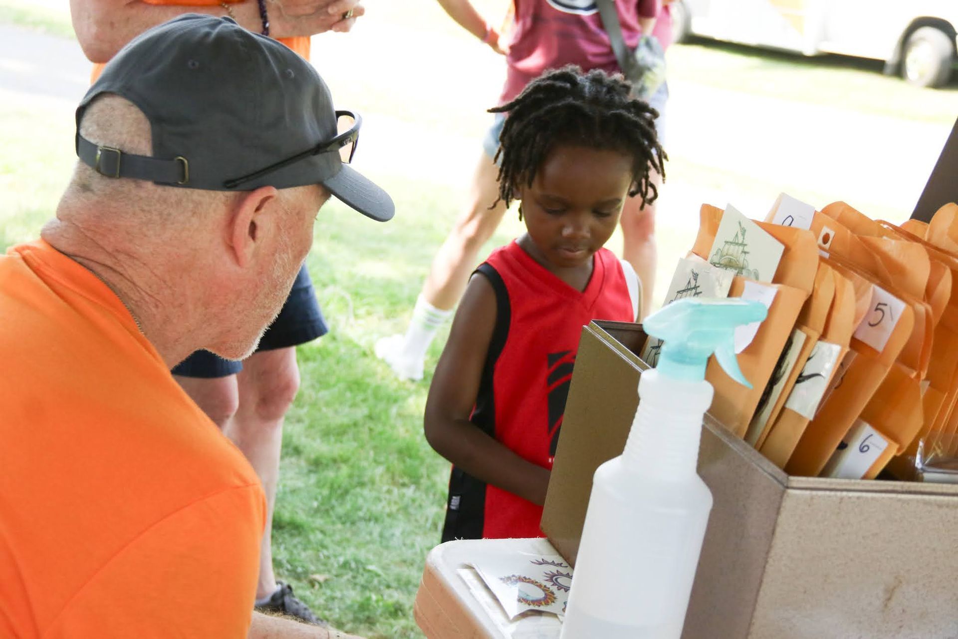 Man in orange shirt assisting a child at a table with seed packets; outdoor setting.