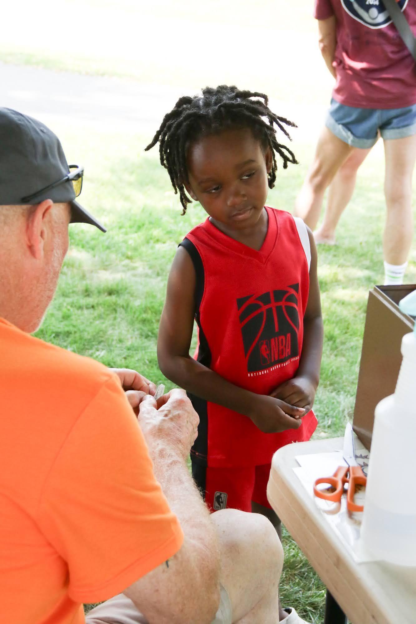 Boy in red jersey watches person at table, outdoor setting.