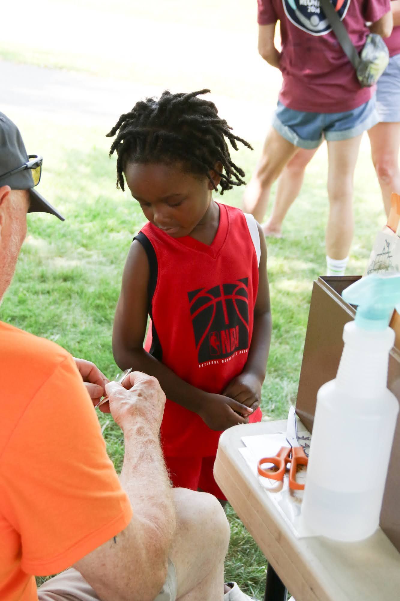 Young person in red jersey watches a person's hand near a table with scissors and spray bottle.
