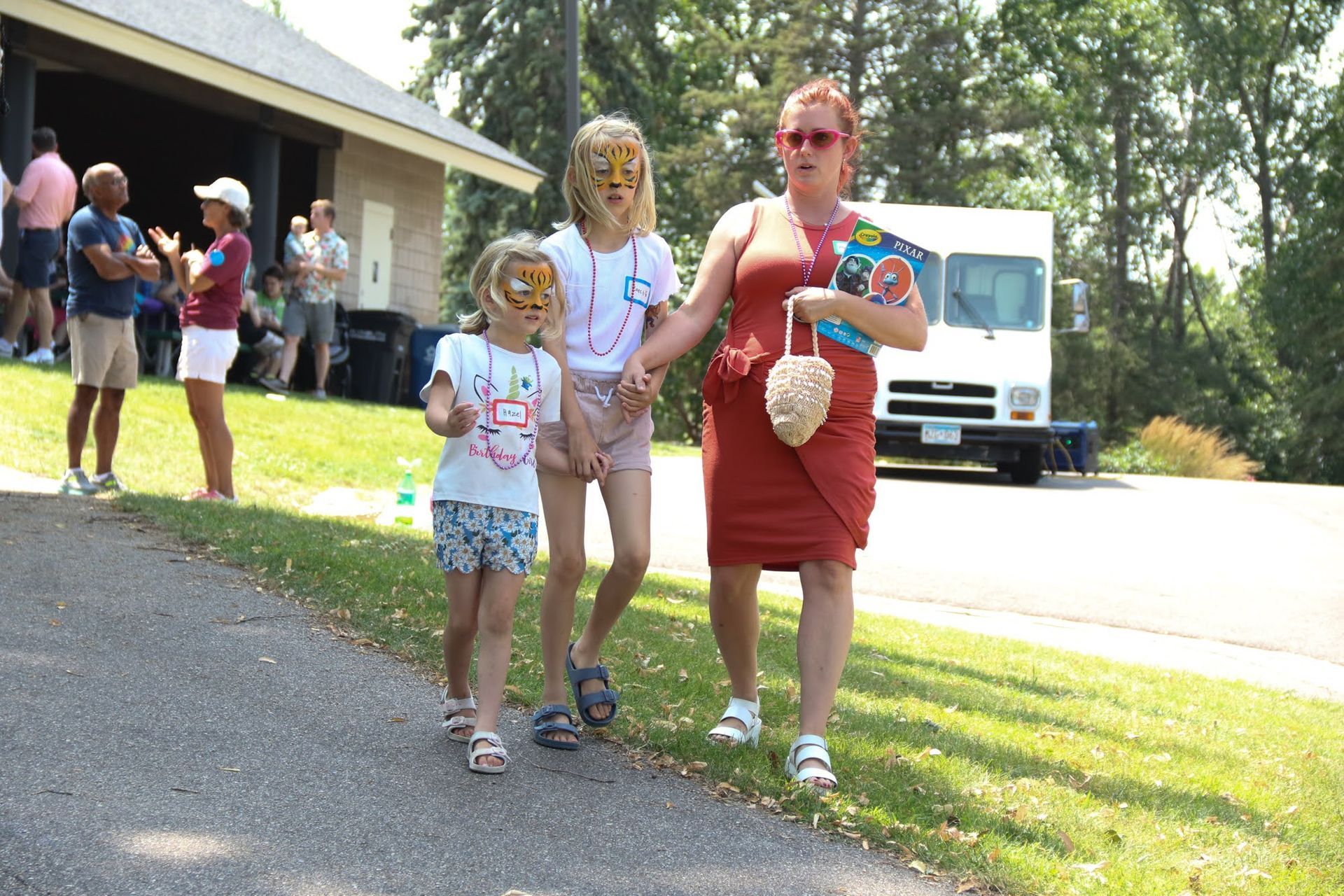 Woman and two children walk on a path, wearing masks. A white truck and people in the background.