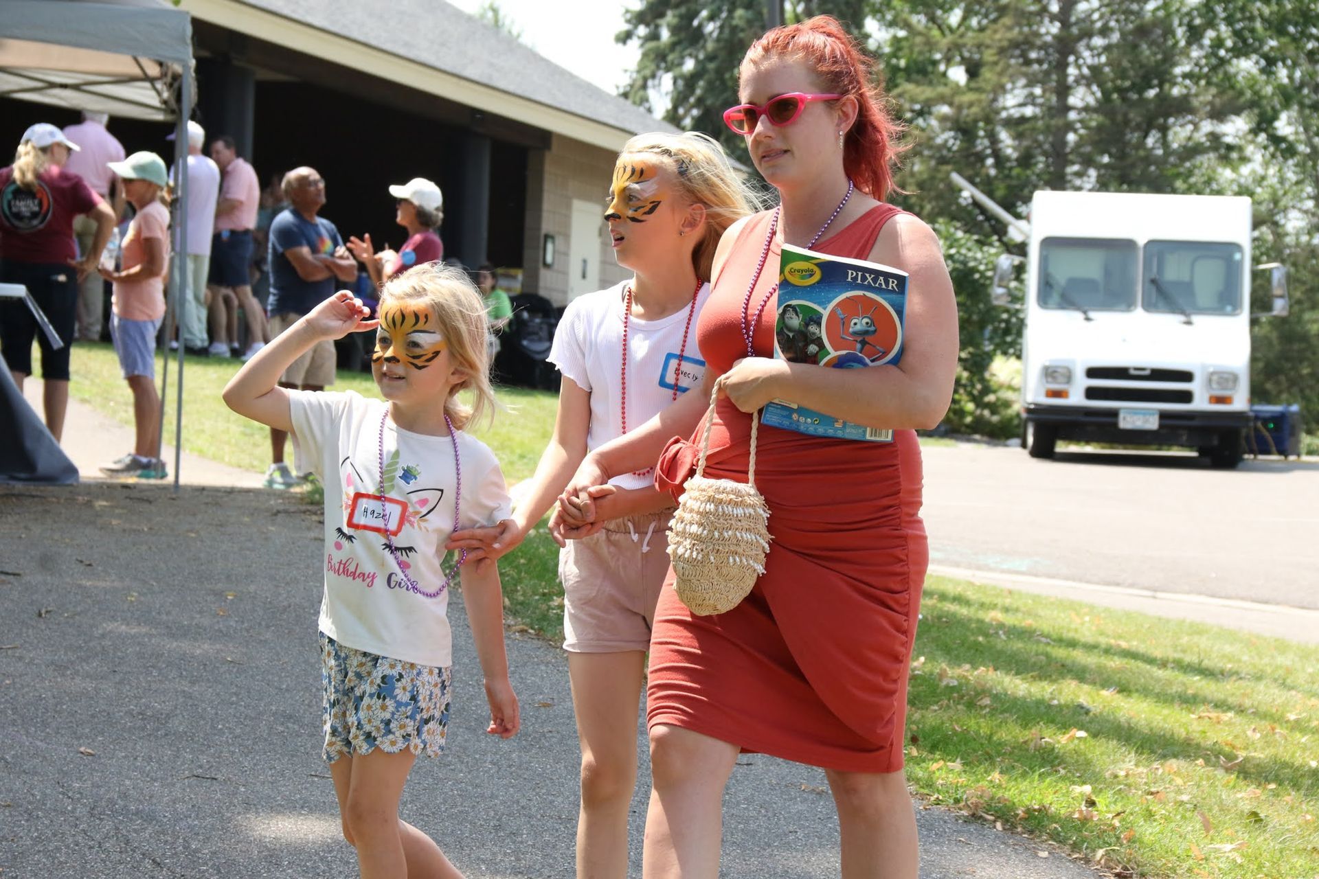 Woman holding a book walks with two children, one with face paint, near a building and truck on a sunny day.