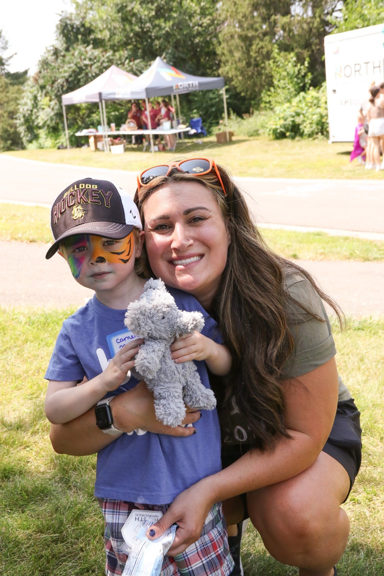 Woman with sunglasses smiles, holding a child with painted face and teddy bear; outdoor event with tents.
