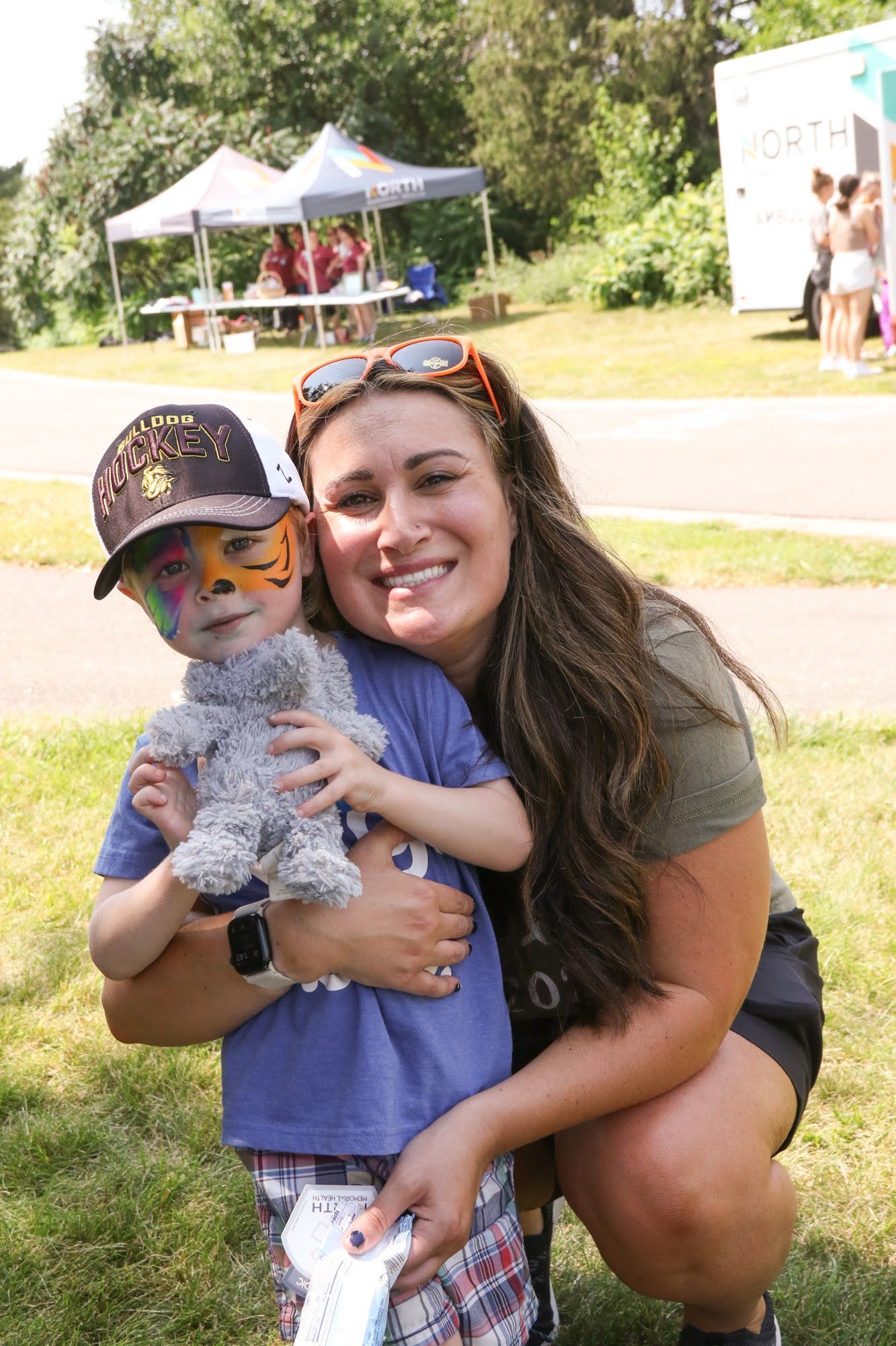 Woman hugs child with face paint holding stuffed animal outdoors.