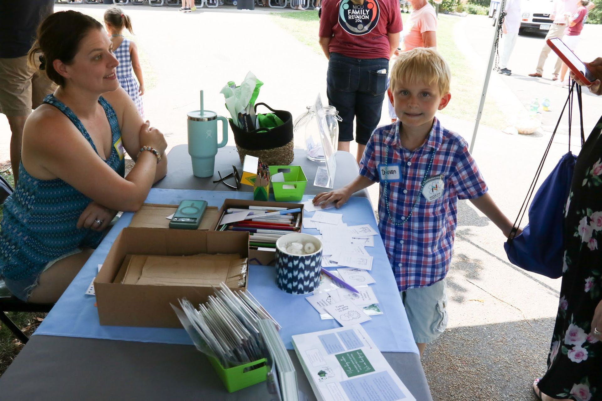 A woman and a young boy stand at a table with papers and supplies outdoors, smiling at the camera.