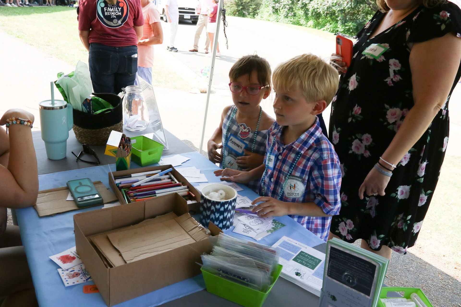 Two children at a table with art supplies; a woman stands nearby. Outdoor setting.