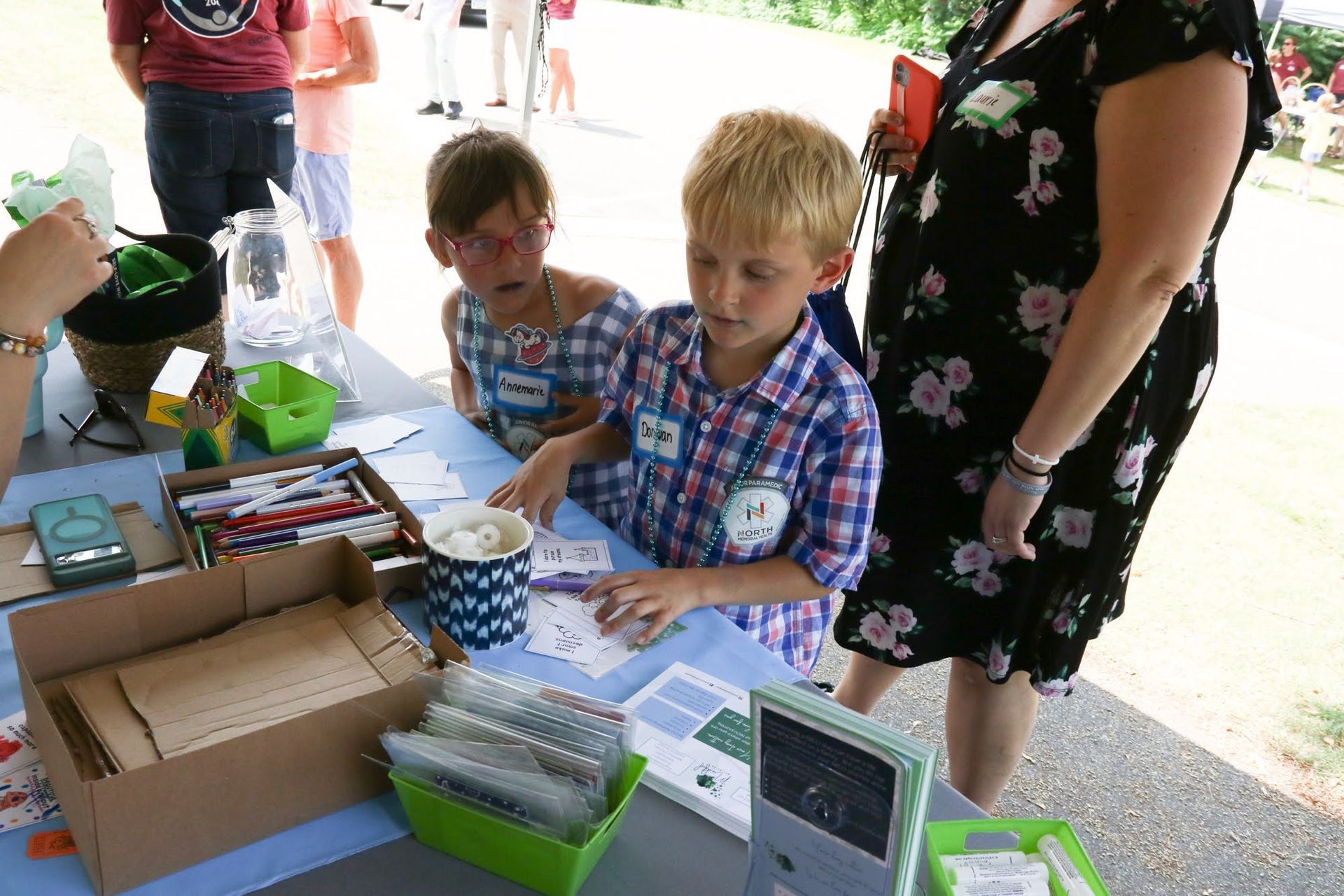 Two children craft at a table outdoors; a woman and other people are nearby.