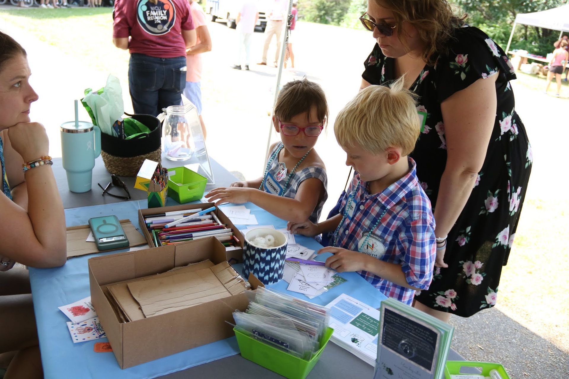 People at a table doing crafts; a woman assists two children; an outdoor setting with a light blue tablecloth.