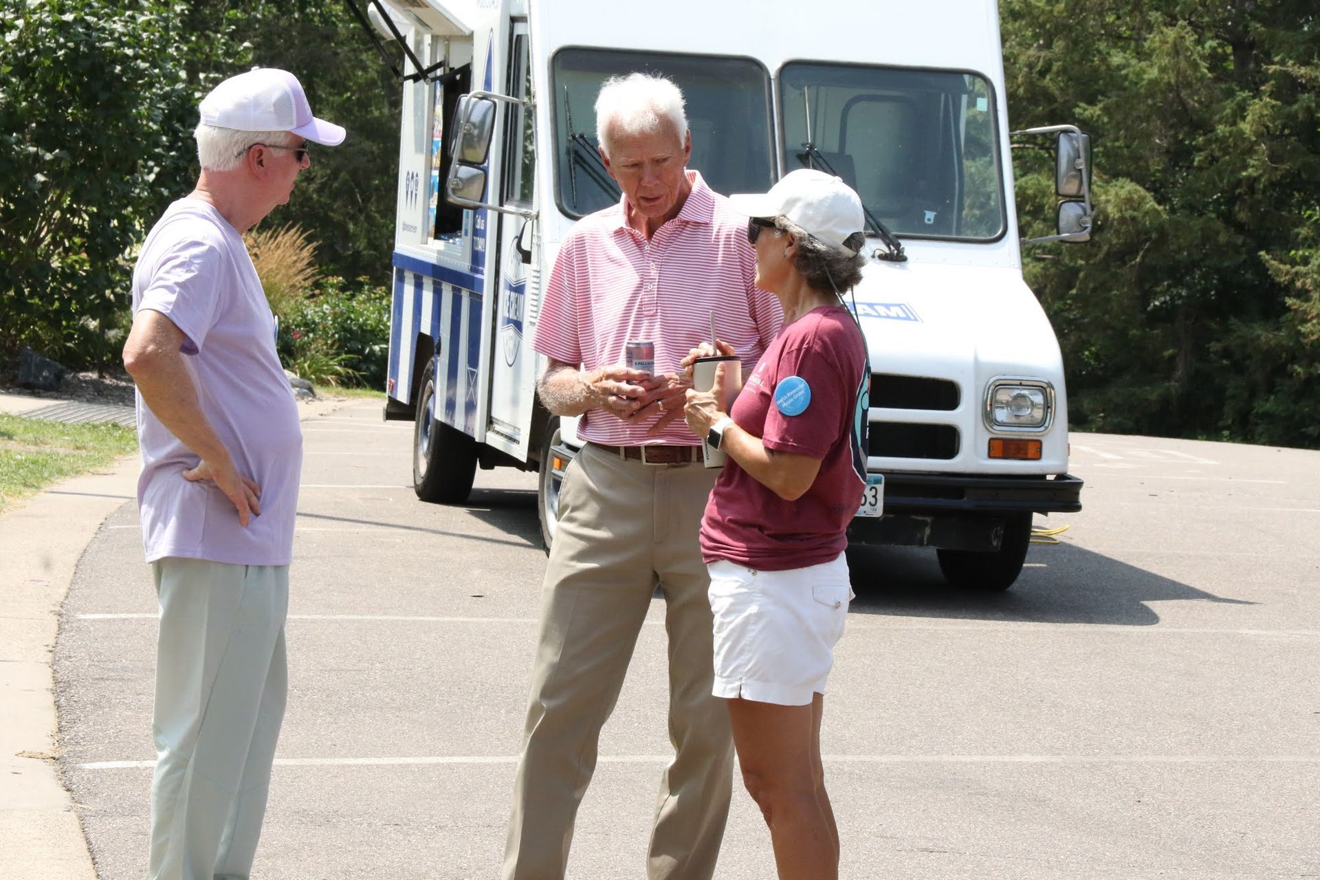 Three people near an ice cream truck. Man in pink shirt shows something to woman in white shorts. Man in purple shirt looks on.