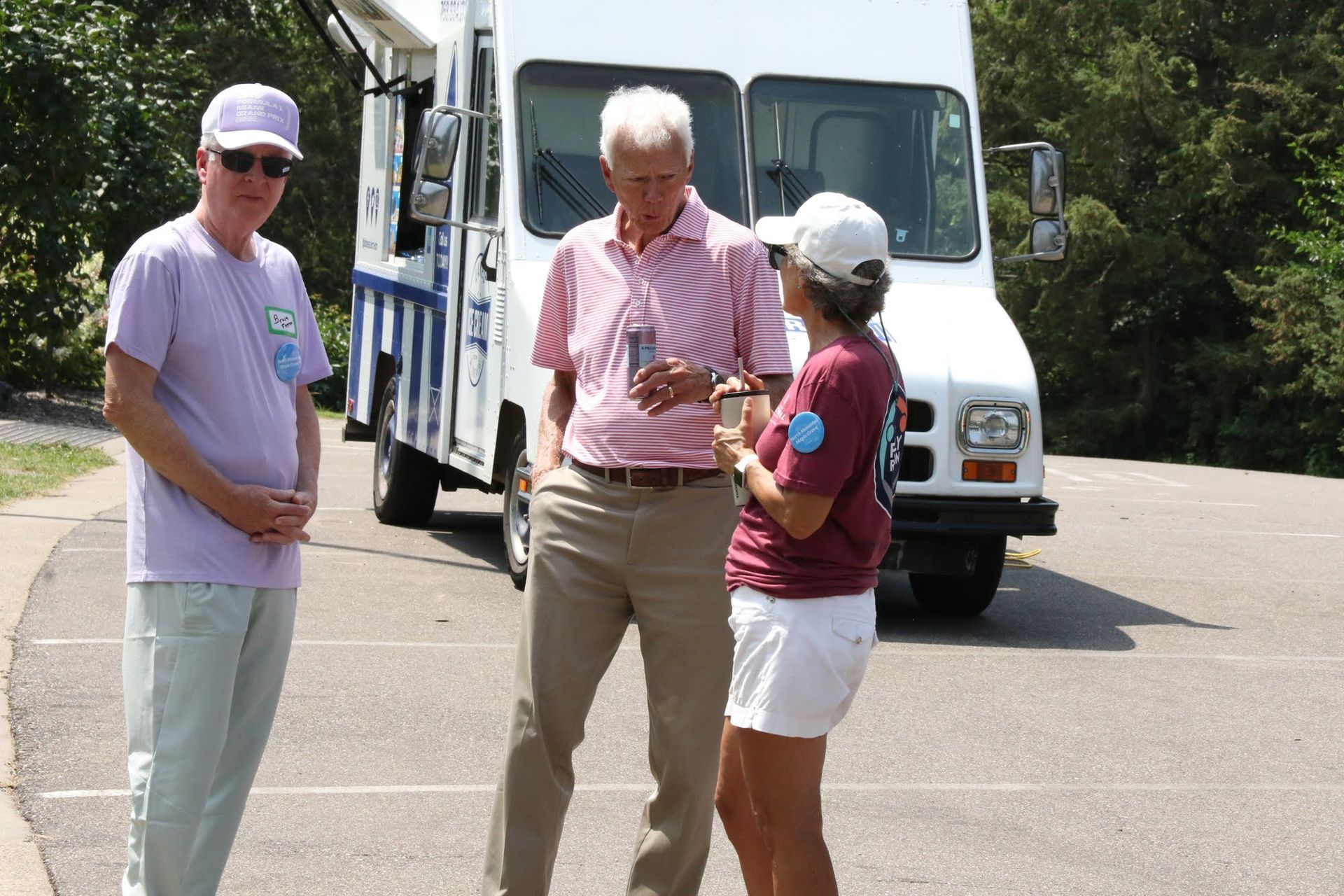 Three people stand near a white truck. Two men and a woman talk in a parking lot on a sunny day.