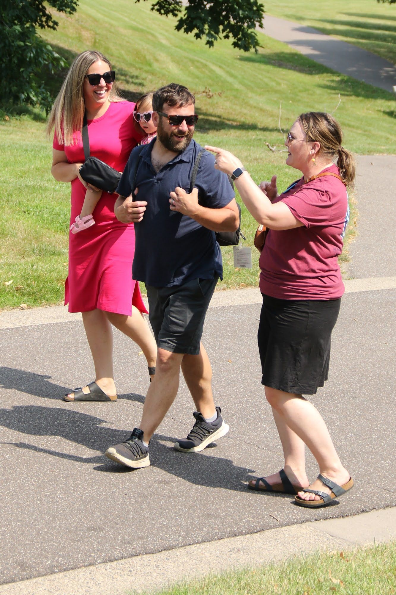 People walking on a paved path outdoors. A woman in pink carries a child, and a man in a navy shirt and sunglasses gestures. Another woman touches the man's arm.