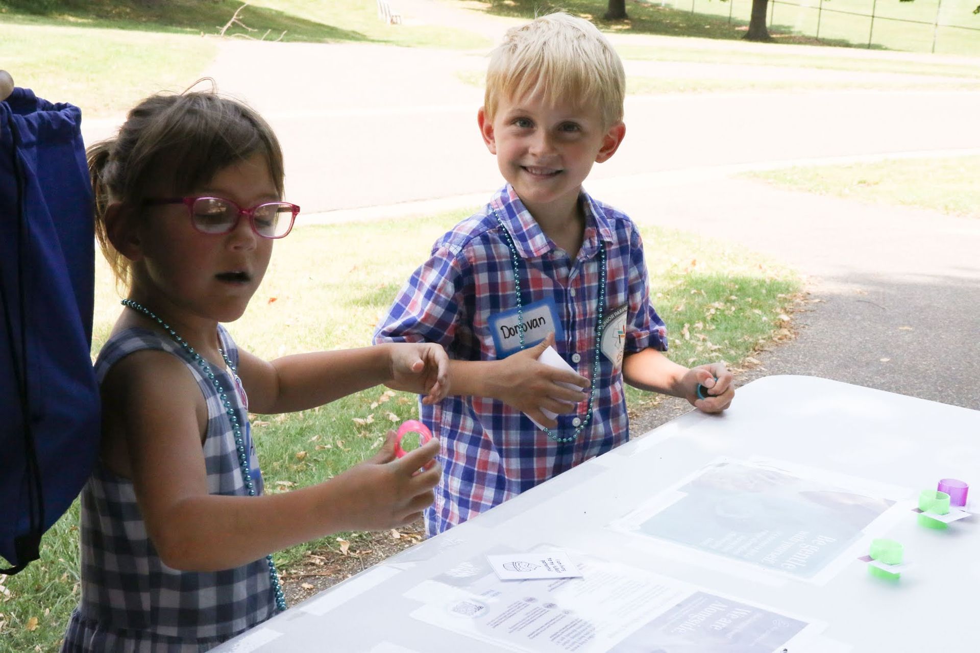 Two children at a table; girl with glasses holding object, boy smiling with name tag; outdoor setting.