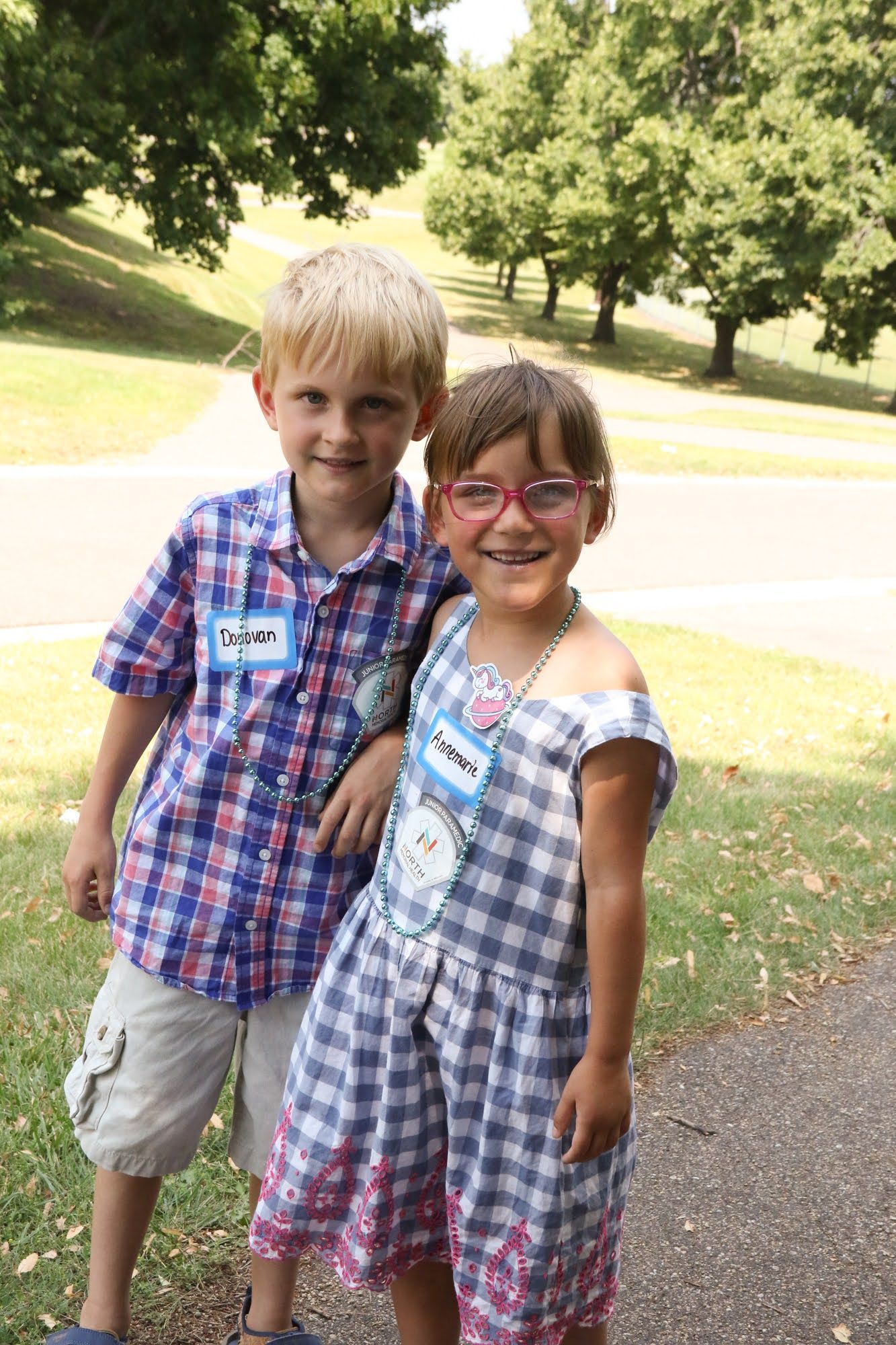 Two children, smiling, stand close together outdoors. Boy wears plaid shirt and shorts; girl, checkered dress, glasses, name tags visible.