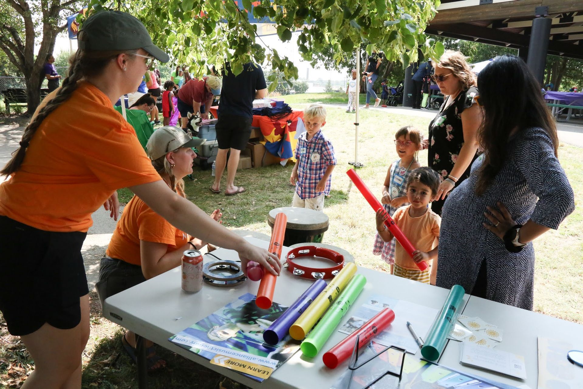 People at an outdoor event with musical instruments at a table.  Children and adults interact with colorful sound makers.