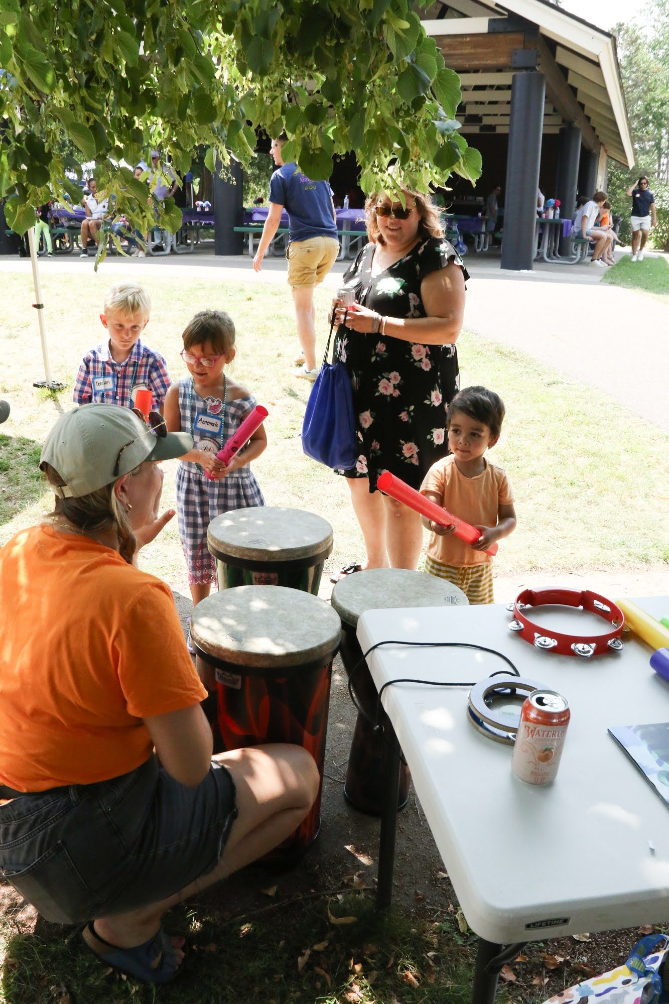 Children playing drums and other percussion instruments outdoors with an instructor and a woman.