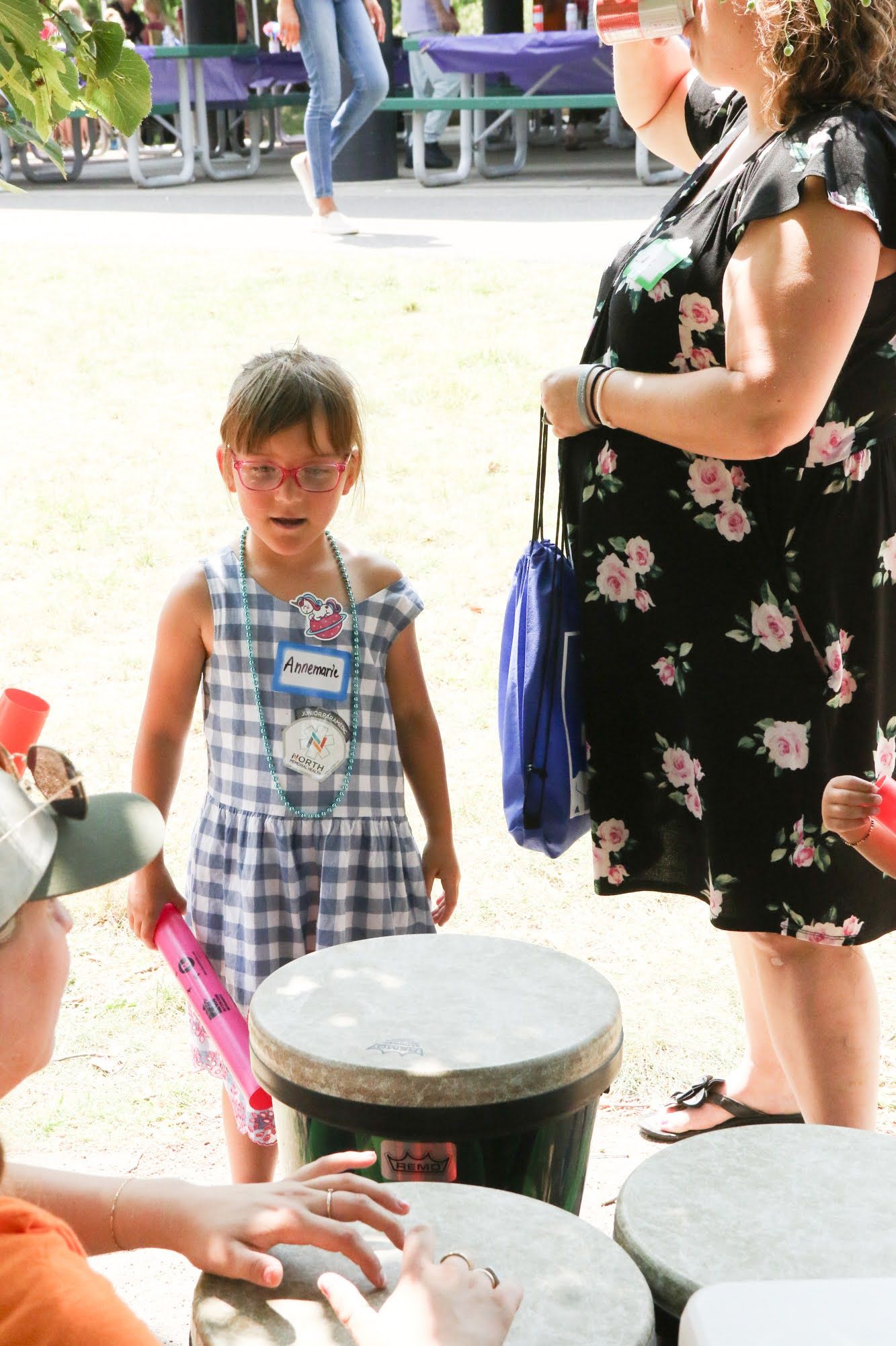 A child in a checkered dress stands near drums, while an adult in a floral dress stands beside them outdoors.