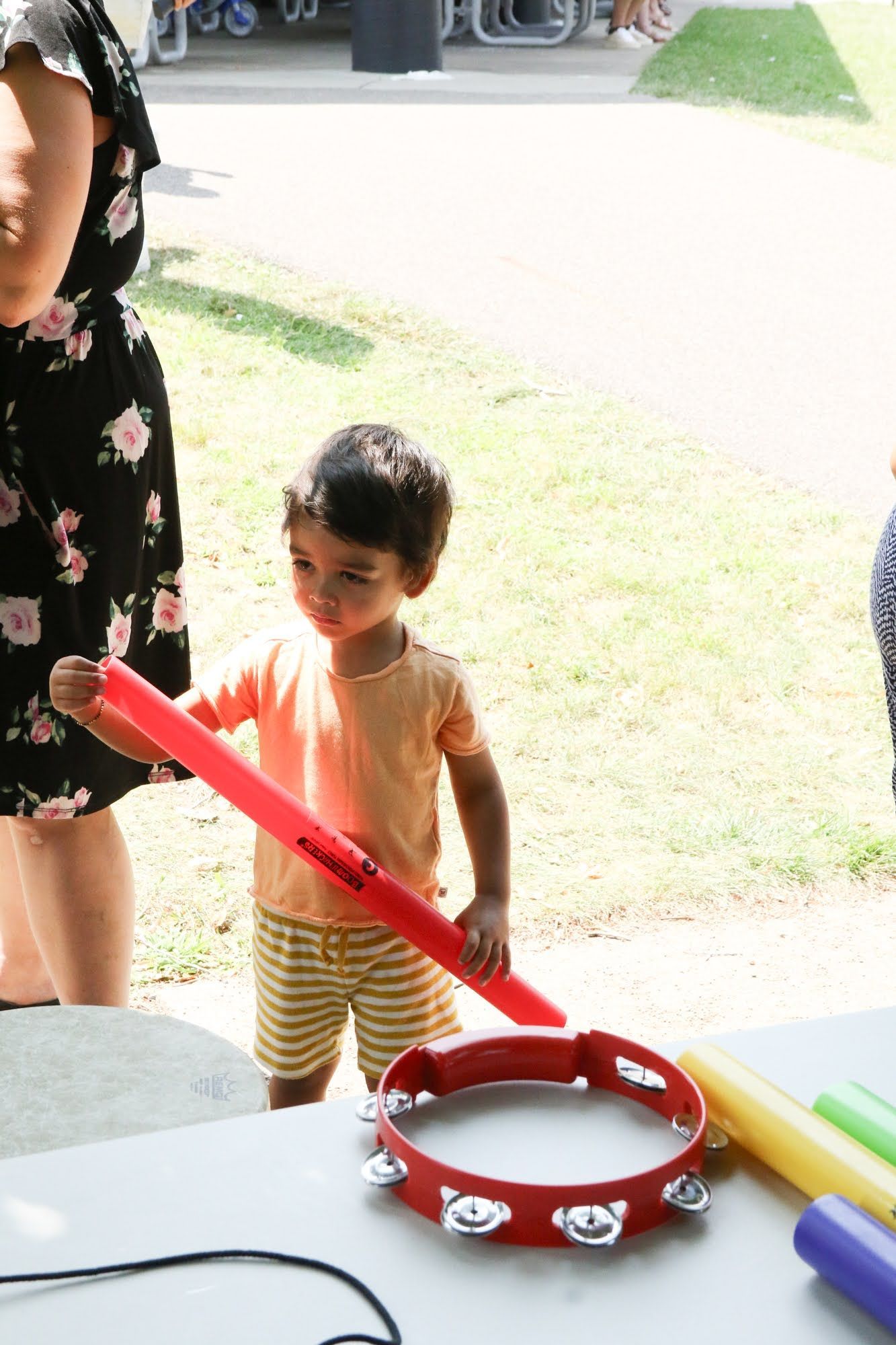 Young child holding red musical stick, looking at tambourine on table outdoors.
