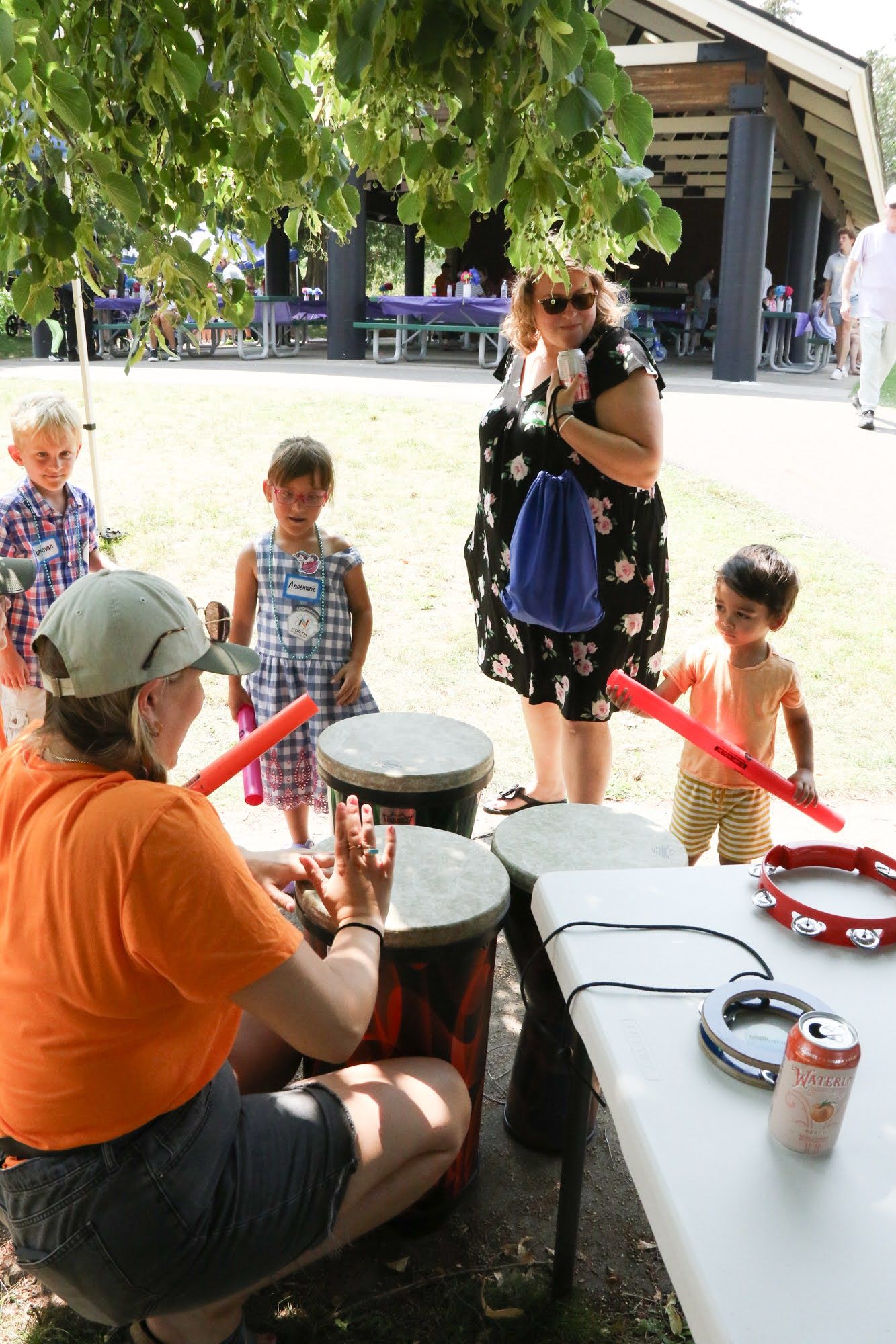 Woman in orange shirt drums for children at outdoor event.