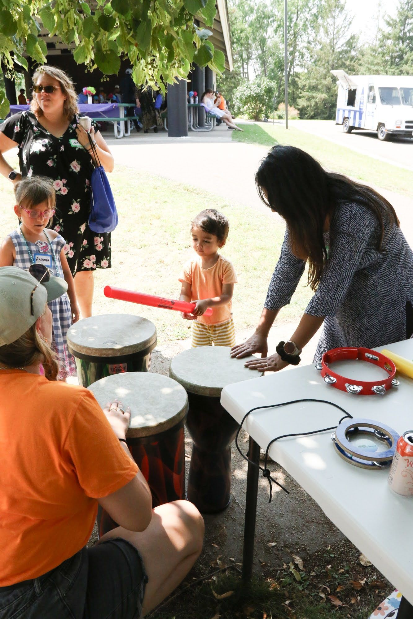 People playing drums at a park. A child holds a drumstick while others look on.