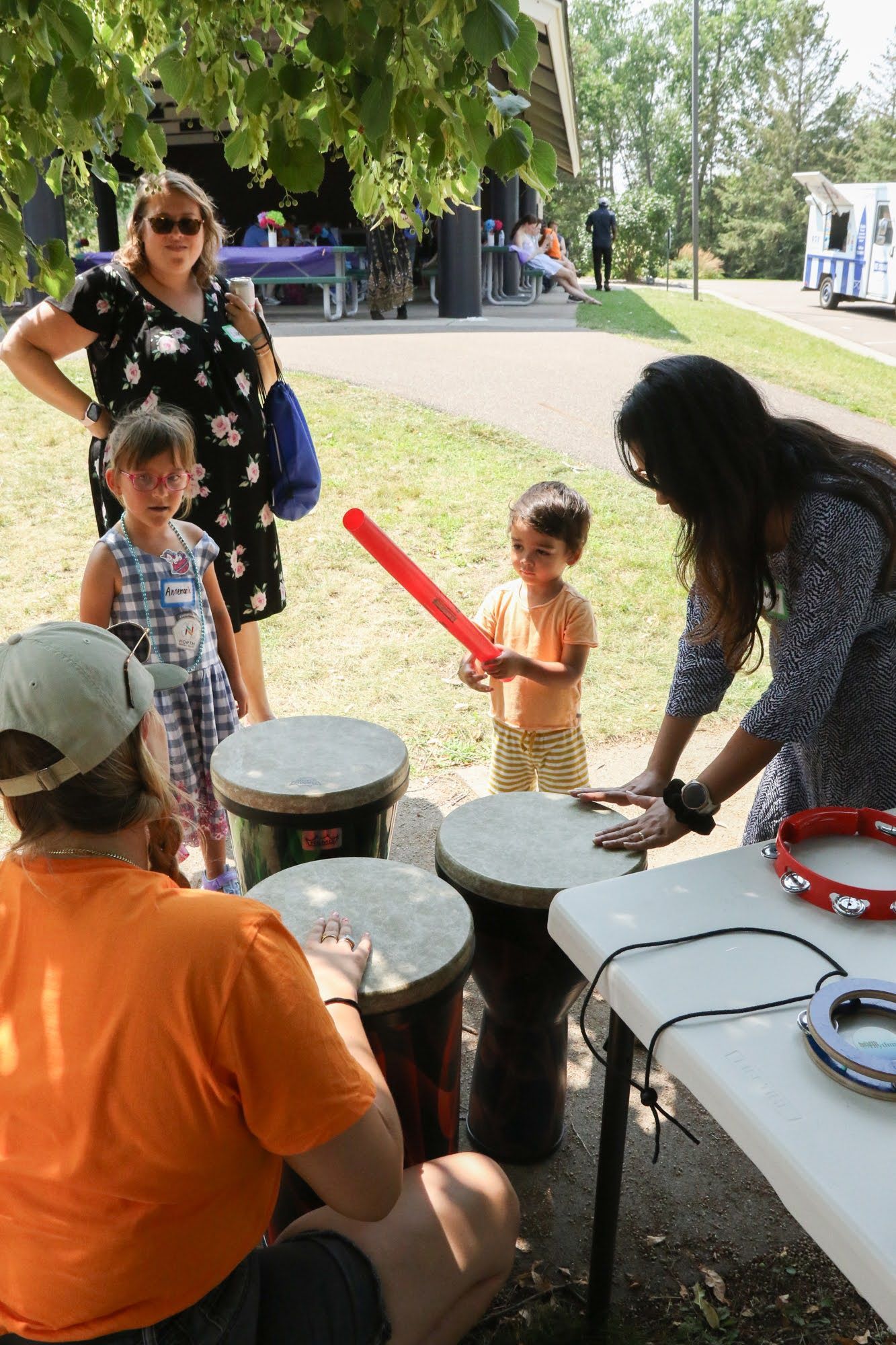 People at a park: a child with a toy sword plays drums, others watch and play instruments.