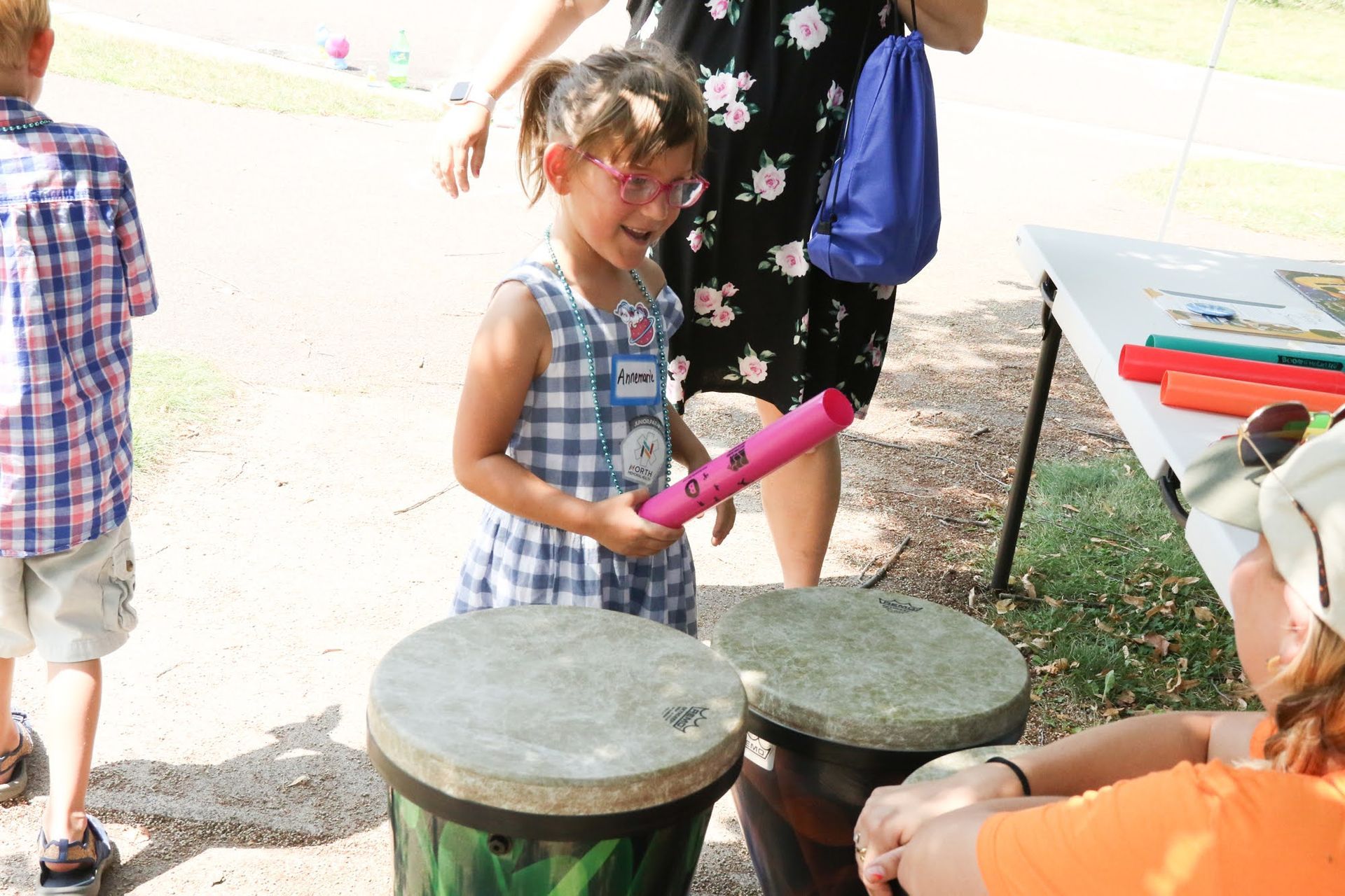 Girl with pink eyewear, holds pink tube, smiles at outdoor drum set.