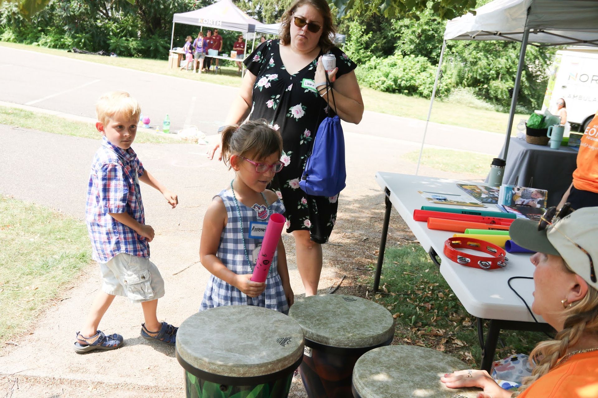 Children playing drums at an outdoor event, attended by an adult with instruments on a table.