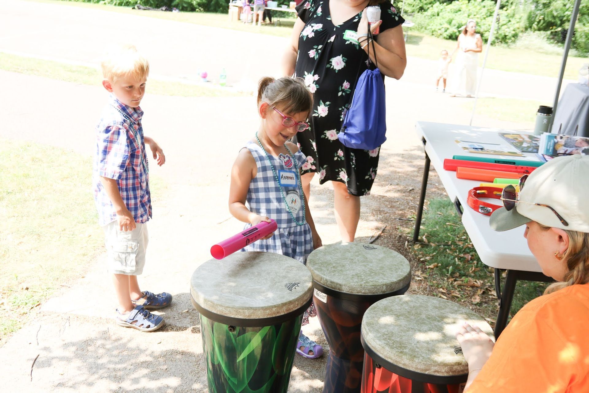 Children playing drums at an outdoor event. One girl smiles and hits a drum with a stick.