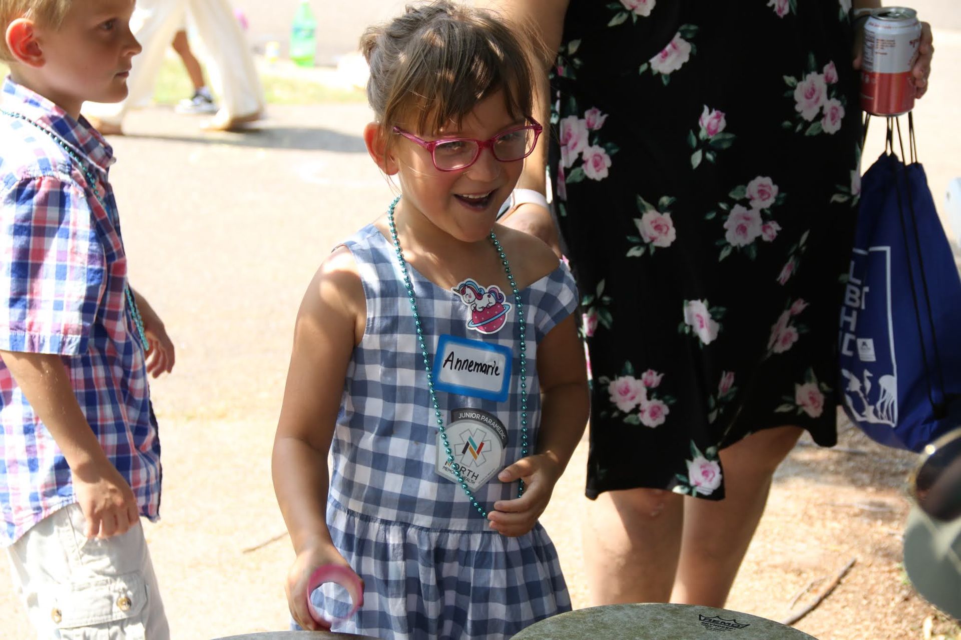 Girl in blue and white dress with pink glasses smiles, a boy next to her and an adult in floral dress.