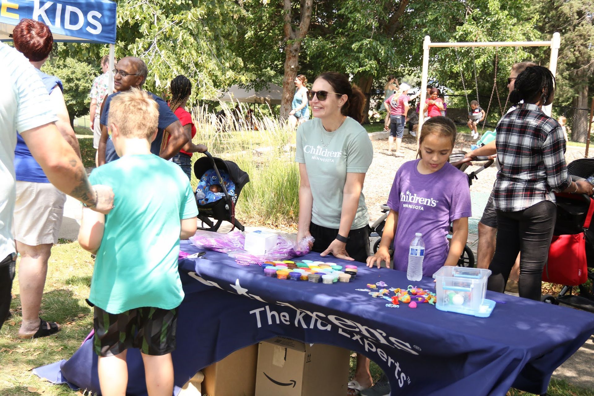 People at an outdoor table with activities. A woman smiles, assisting children. Other people stroll in a park setting.