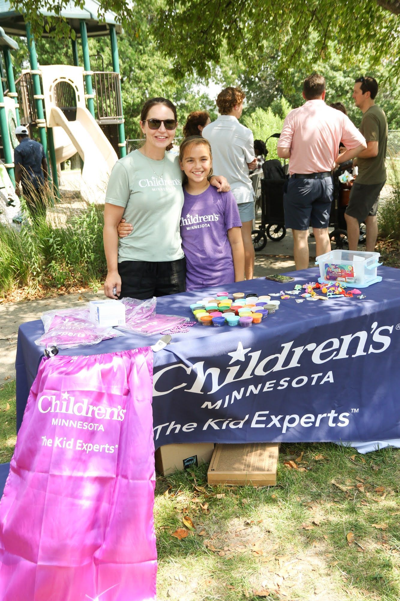 Woman and child at a Children's Minnesota booth.  They are smiling.  Table with purple and blue signage, toys, and giveaways.