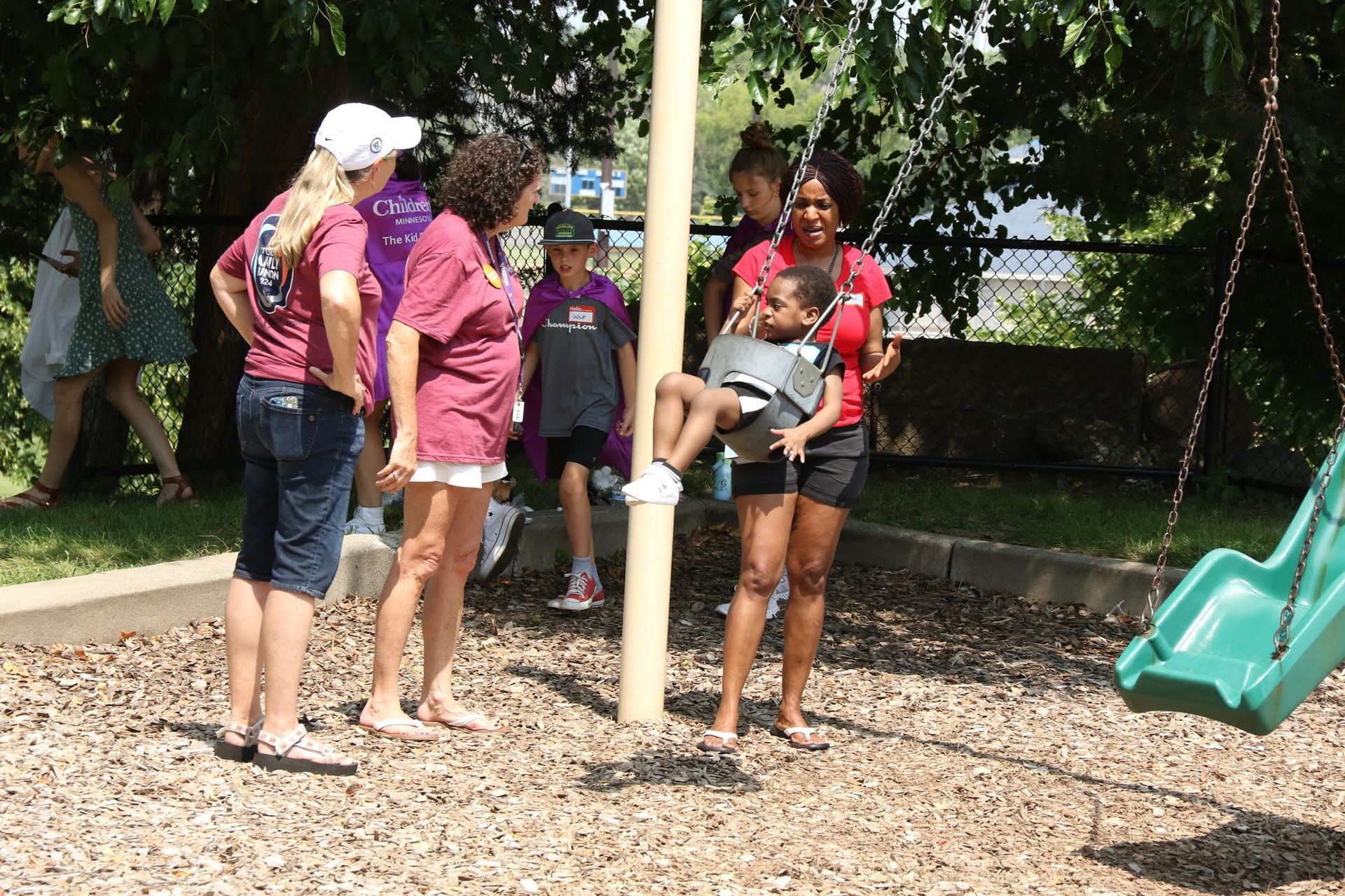 People at a playground, one holding a child near a pole, others watch. Wooden chips ground cover, green slide.