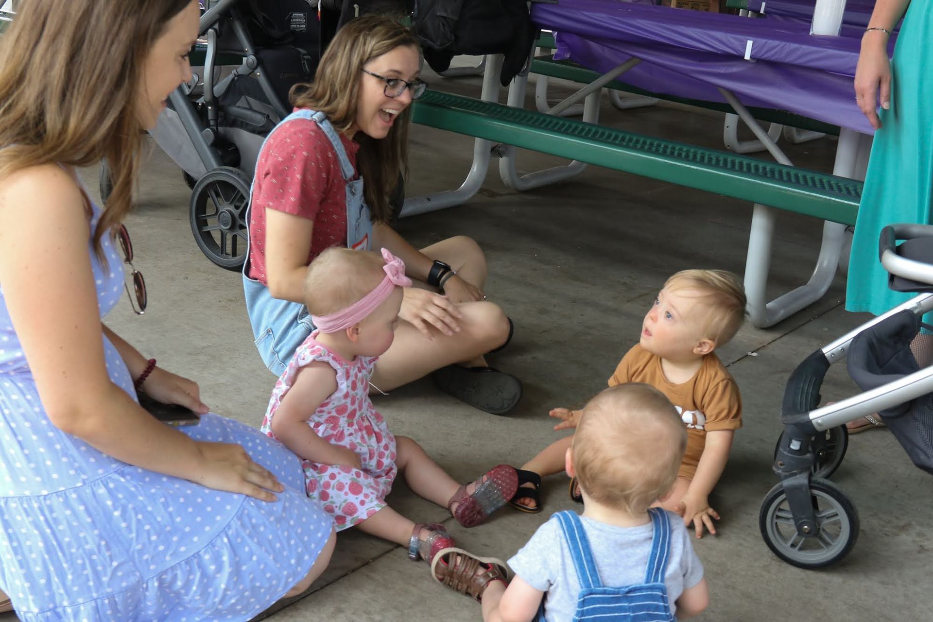 Four babies sit on the ground, interacting with two women at an outdoor gathering.