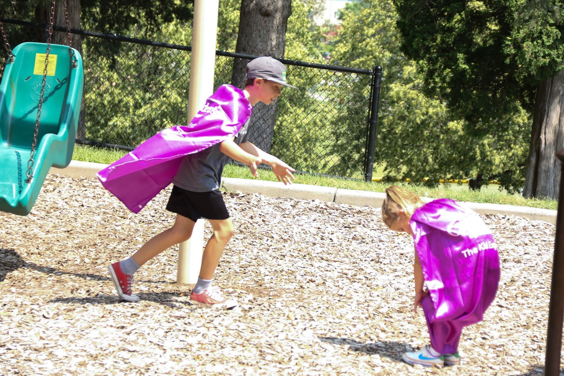 Two children in purple capes play outside by a slide on wood chips.