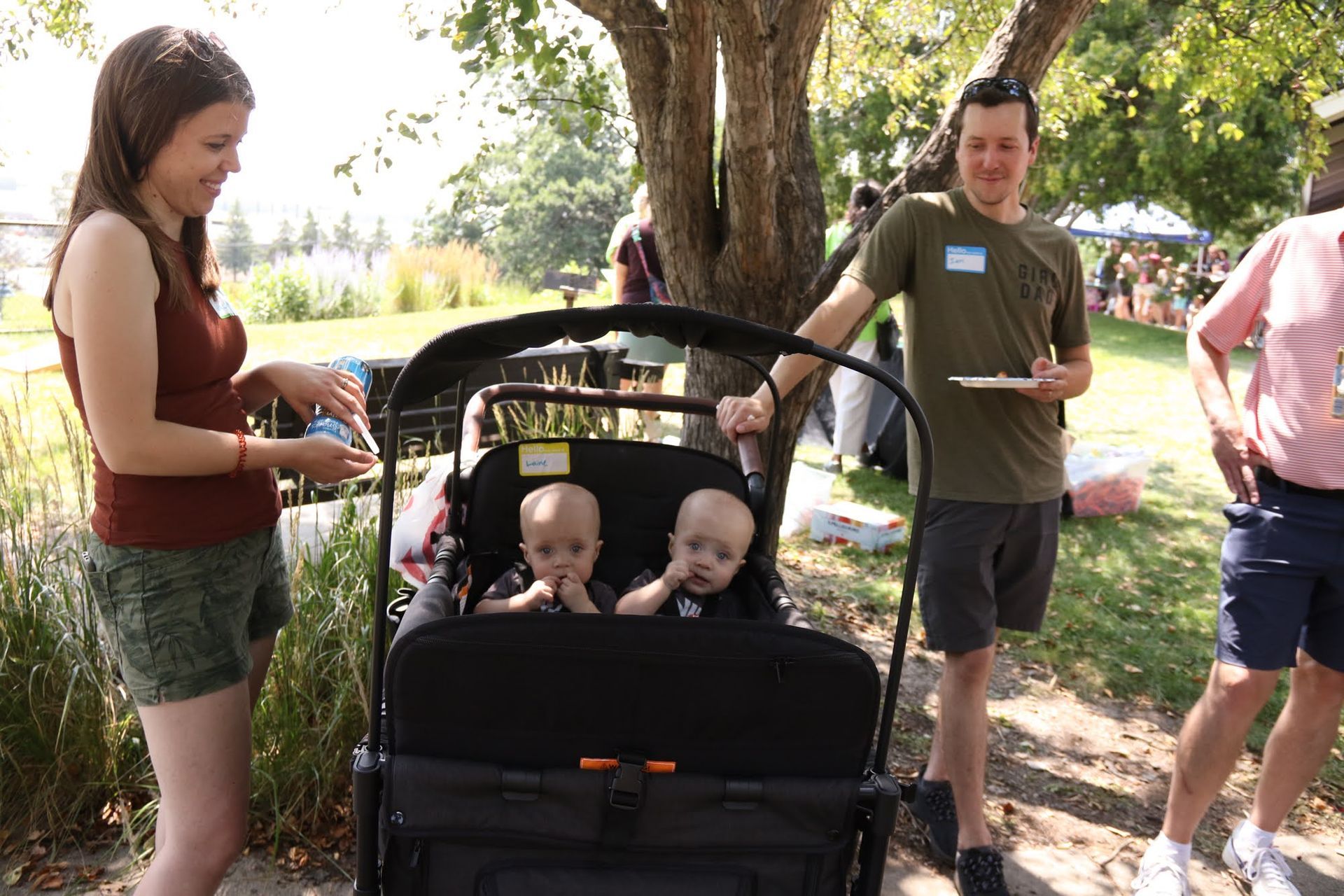 Two babies sit in a stroller. Woman and man stand near them outside on a sunny day.