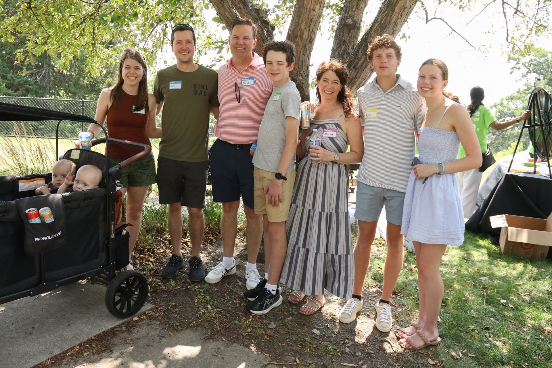 Family posing for photo outdoors near a wagon with babies. Several adults and teens smiling. Sunny day.