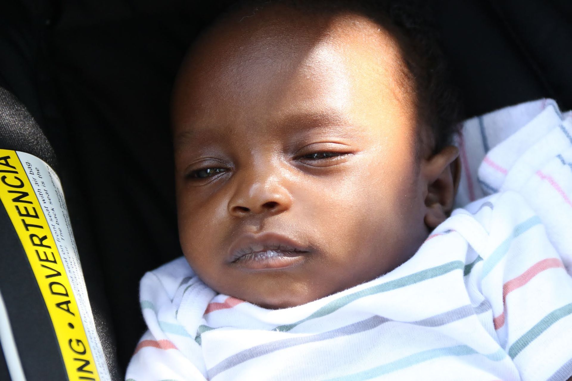 Baby in a white and blue striped onesie, eyes partially closed, in a black stroller.
