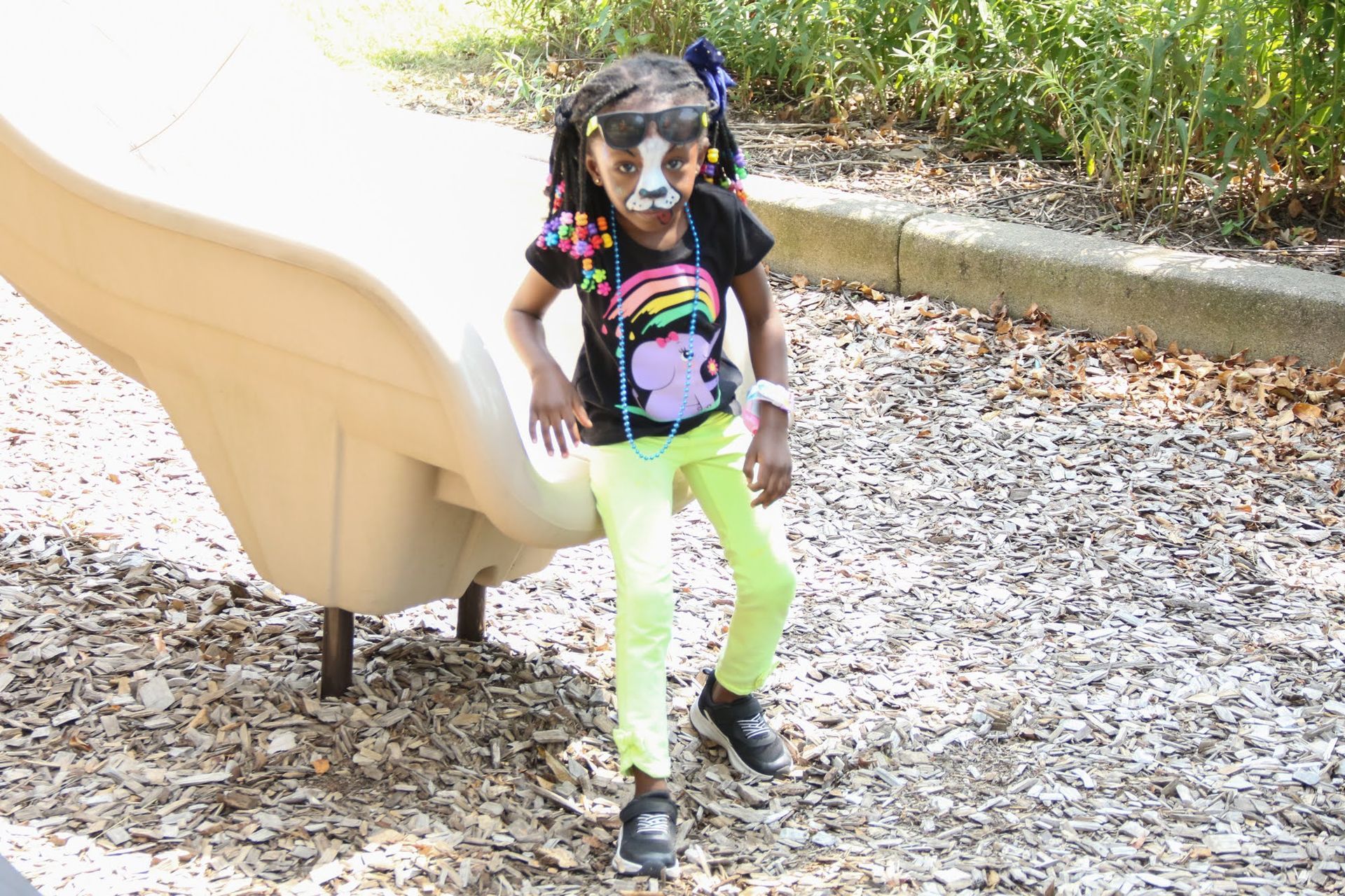Child with face paint coming down a playground slide wearing a rainbow t-shirt and neon pants.