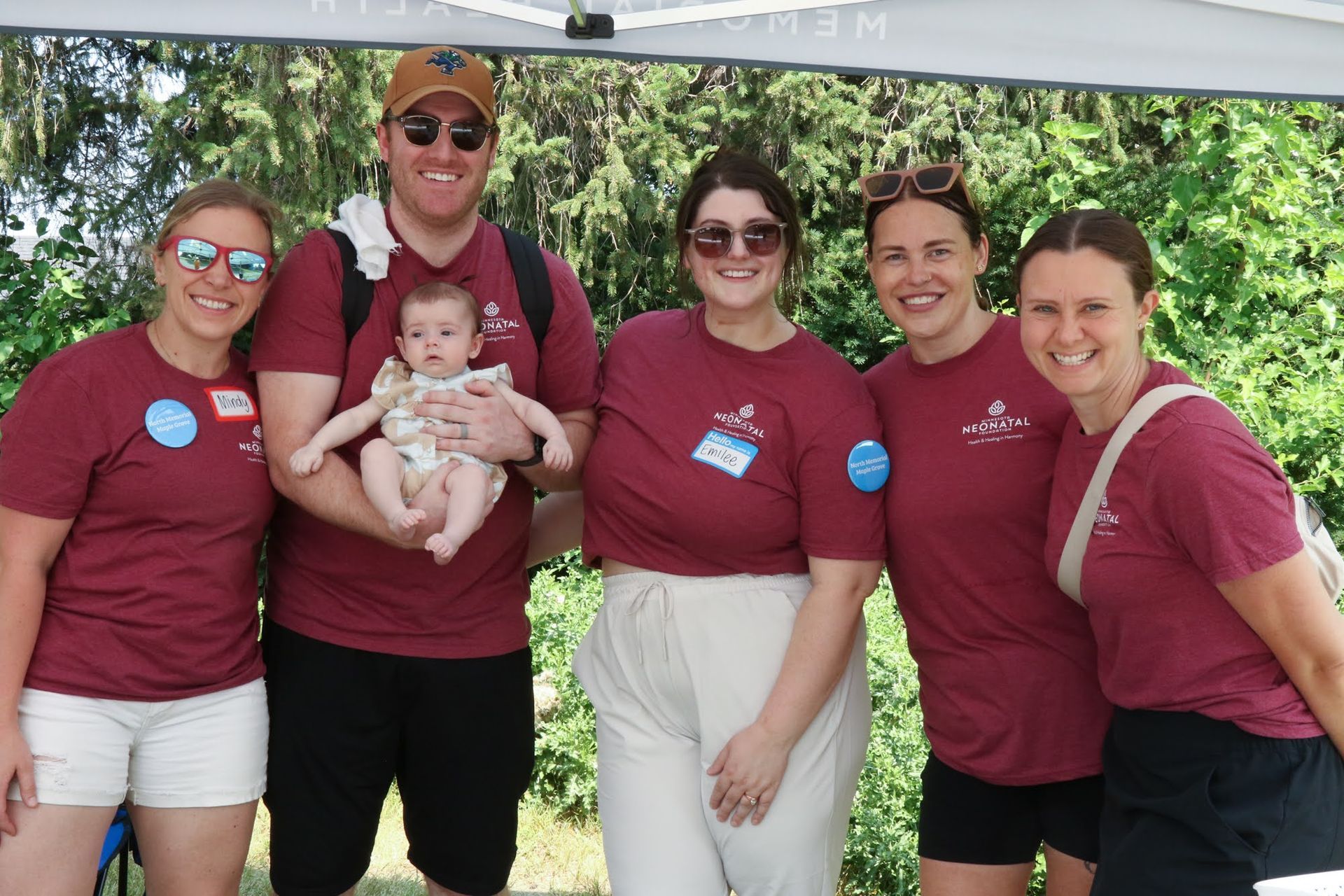 Group of six smiling people outdoors; one holding a baby, all wearing maroon shirts.