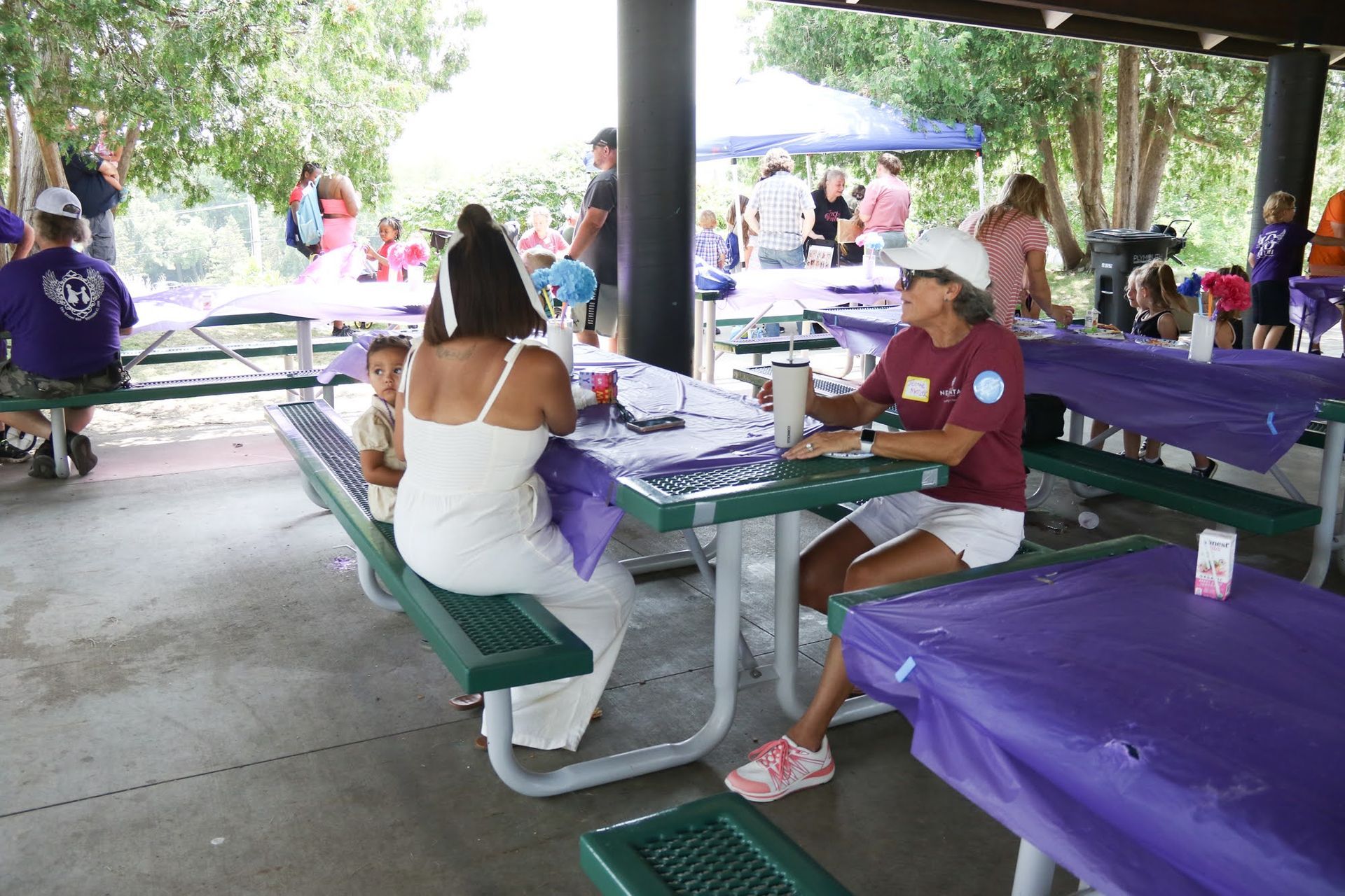 Picnic gathering under a shelter with purple tablecloths and people sitting at tables.