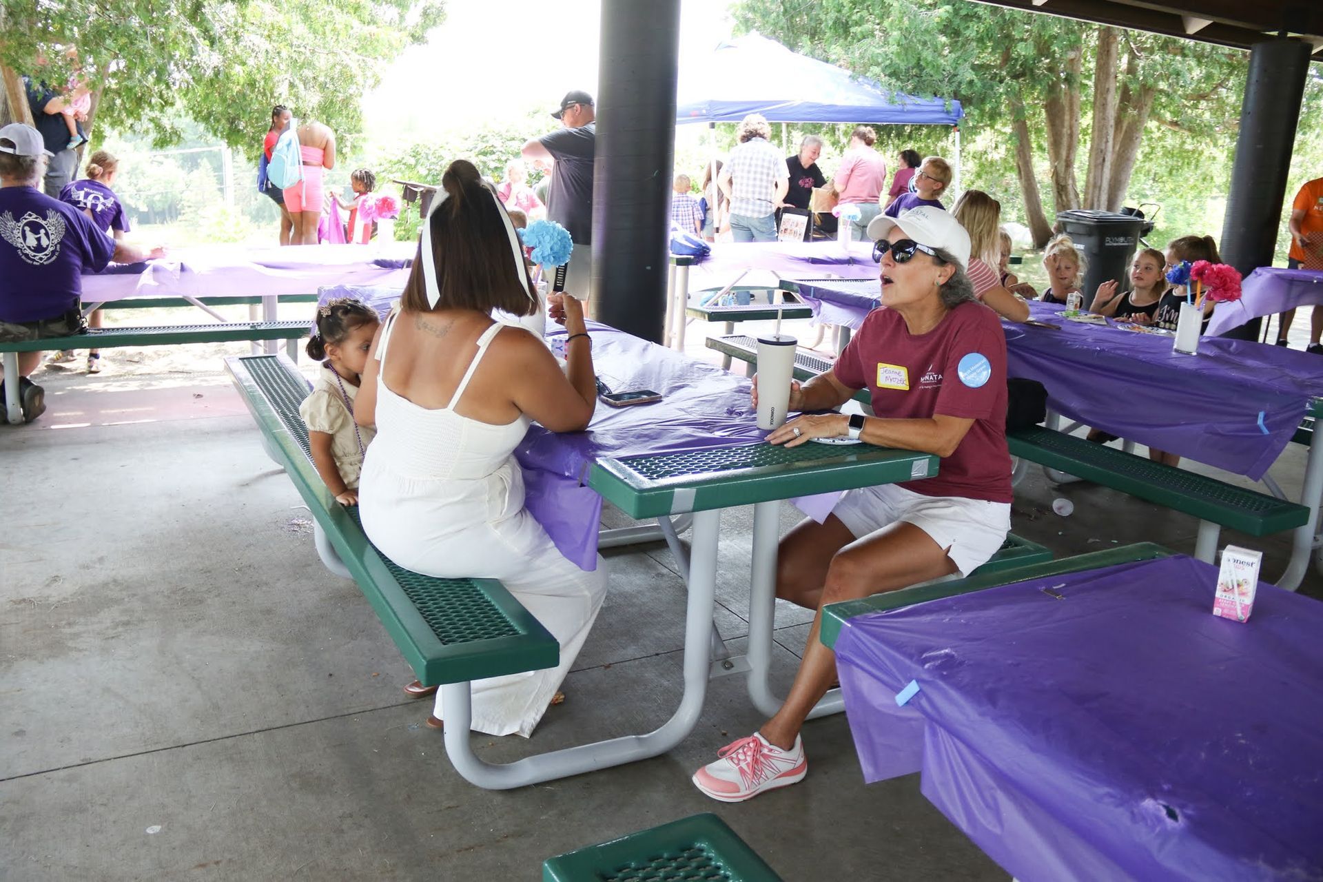 People at picnic tables, one woman in white dress talking to another. Tables covered in purple, outdoor setting.