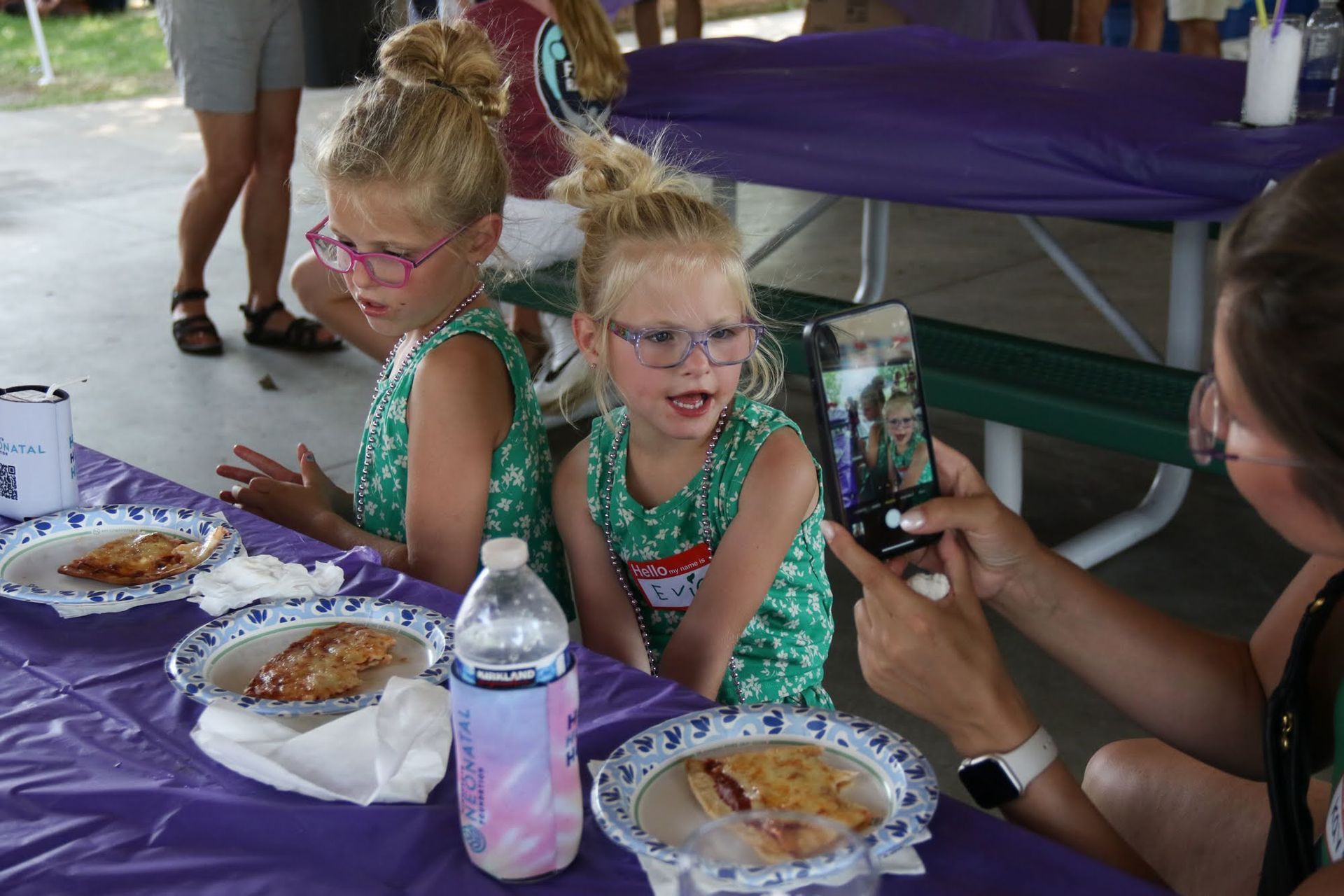 Two young girls with up-do hairstyles, glasses, and matching green dresses sit at a purple table while someone takes a photo.