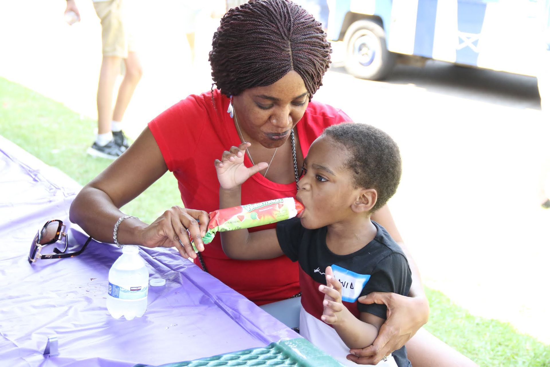 Woman helping a child eat a popsicle at a picnic. Red shirt, watermelon-flavored, outdoor setting.
