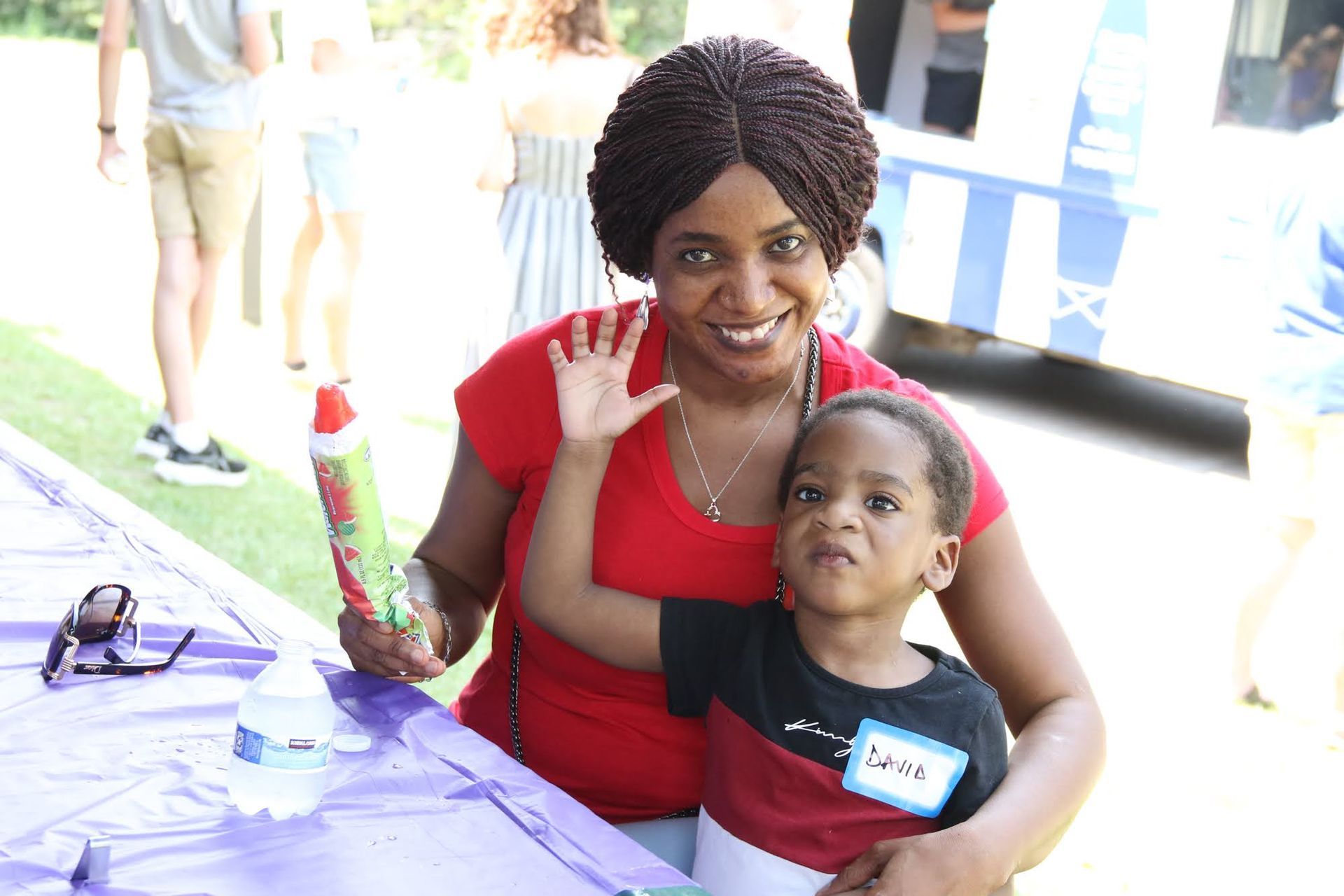 Woman in red top with child, both smiling, at outdoor event; woman waving.