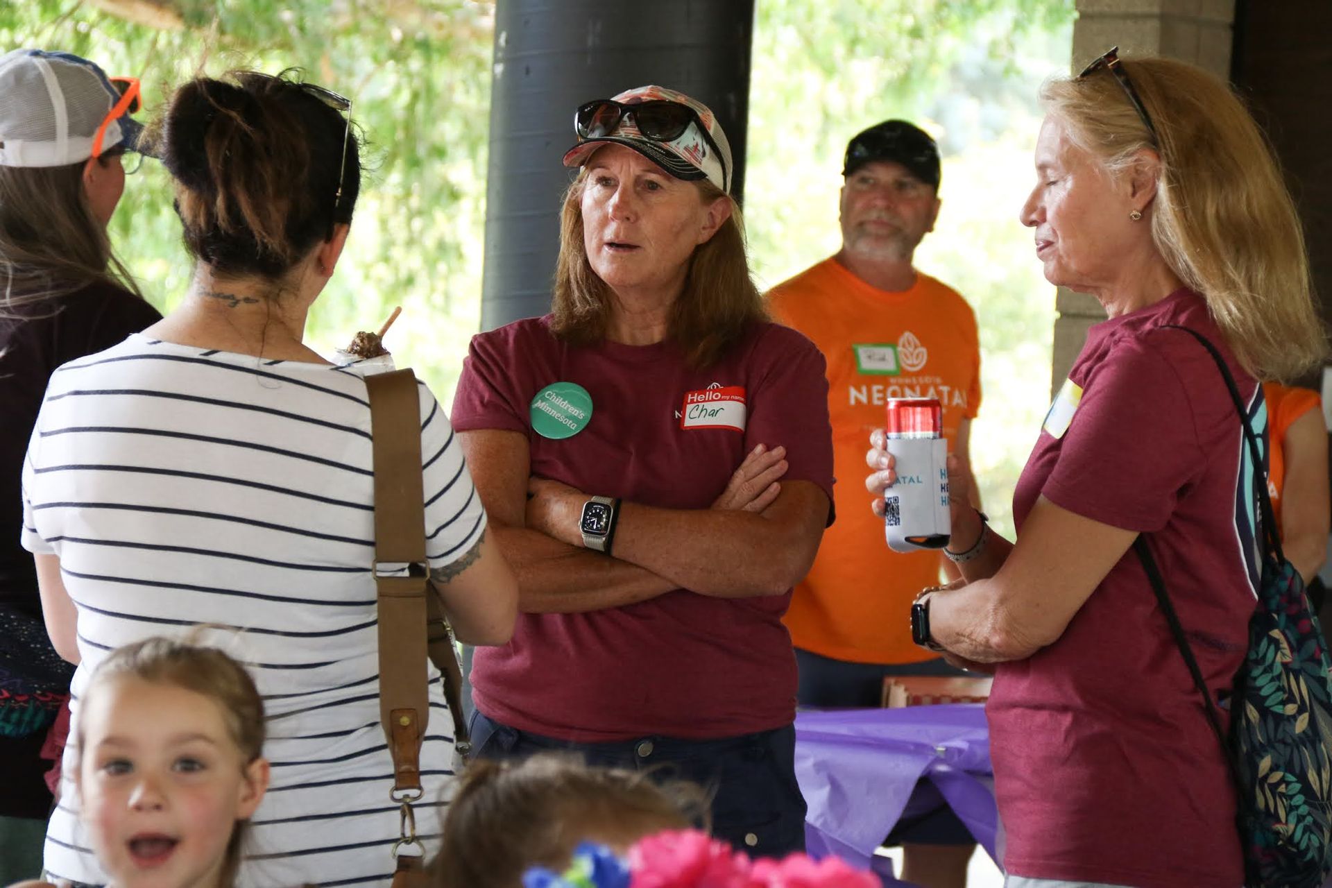 People talking outdoors at an event, wearing casual clothes. One woman has arms crossed, another holds a phone.