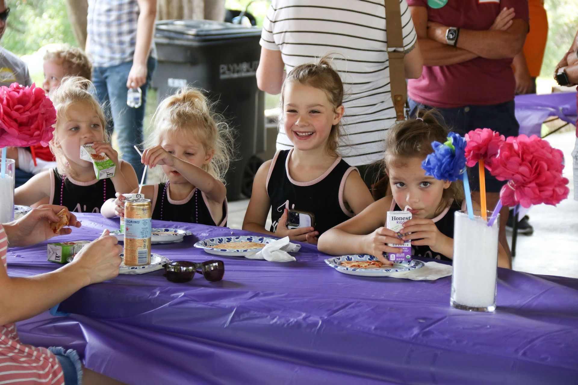 Four young girls at a purple table, eating snacks. One smiles, others drink from juice boxes, with decorative flowers.