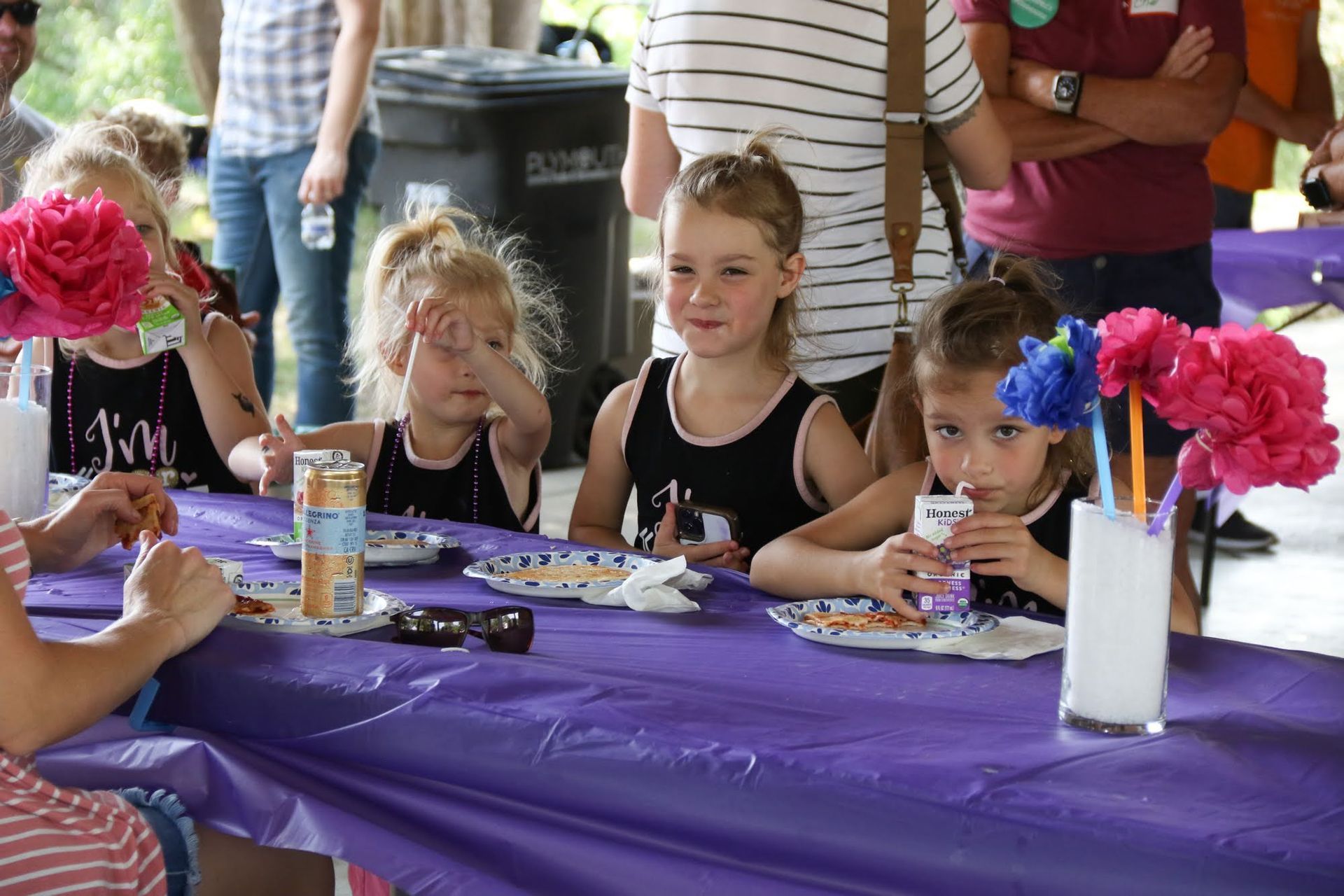 Four young girls sitting at a purple picnic table, with snacks, drinking, and paper flowers.