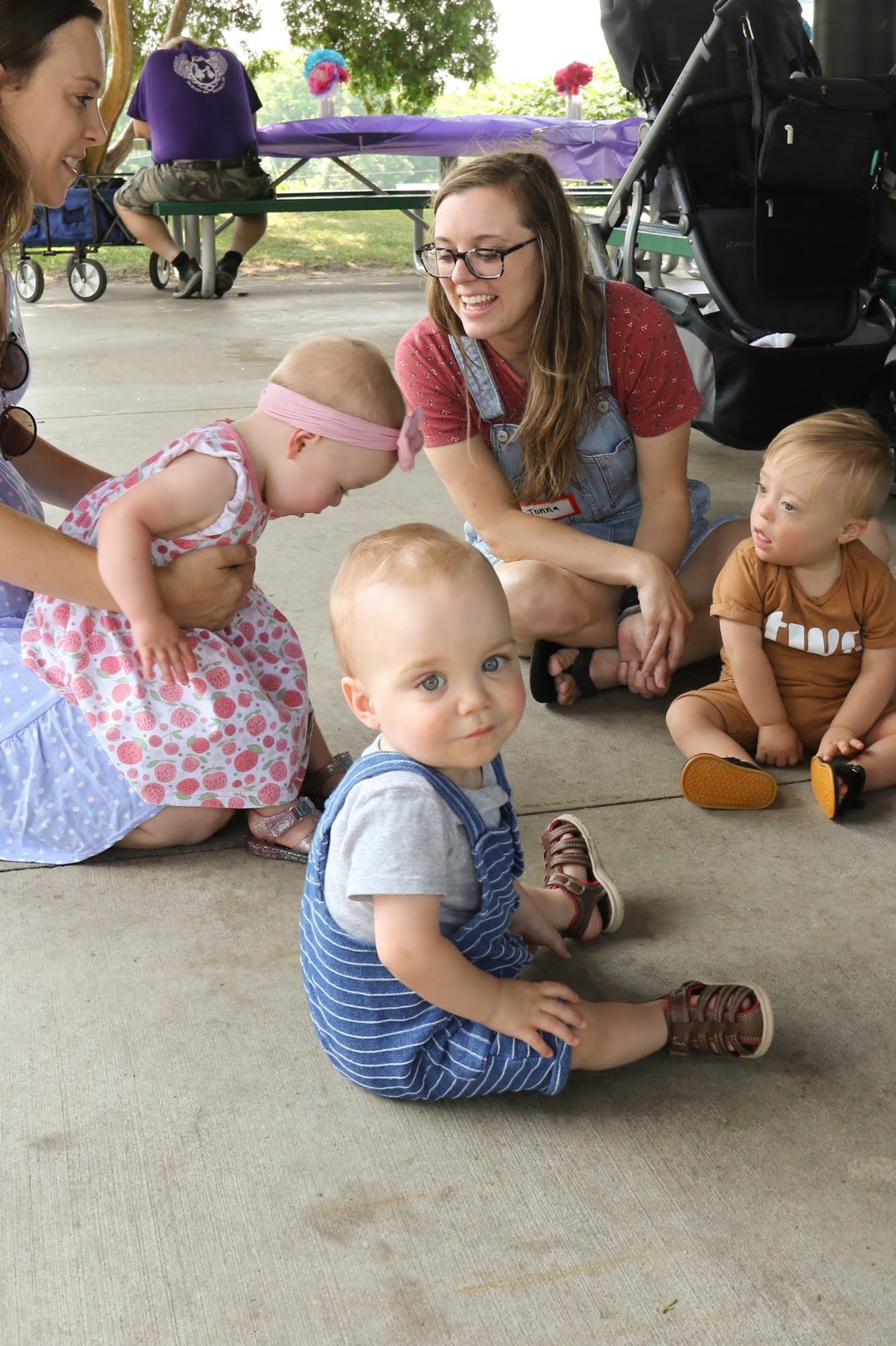 Four babies and two women at outdoor picnic. Babies are sitting on the ground, one looks at the camera.