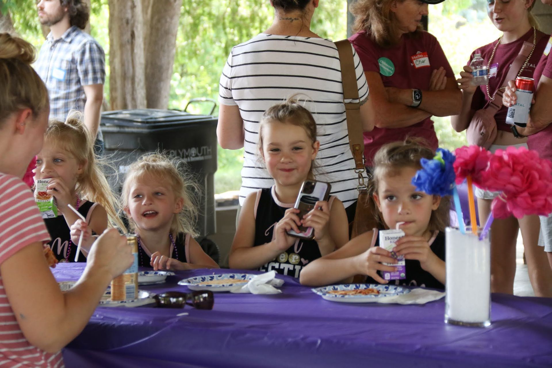 Children at a table with adults nearby; drinking, eating, purple tablecloth, outdoor setting.