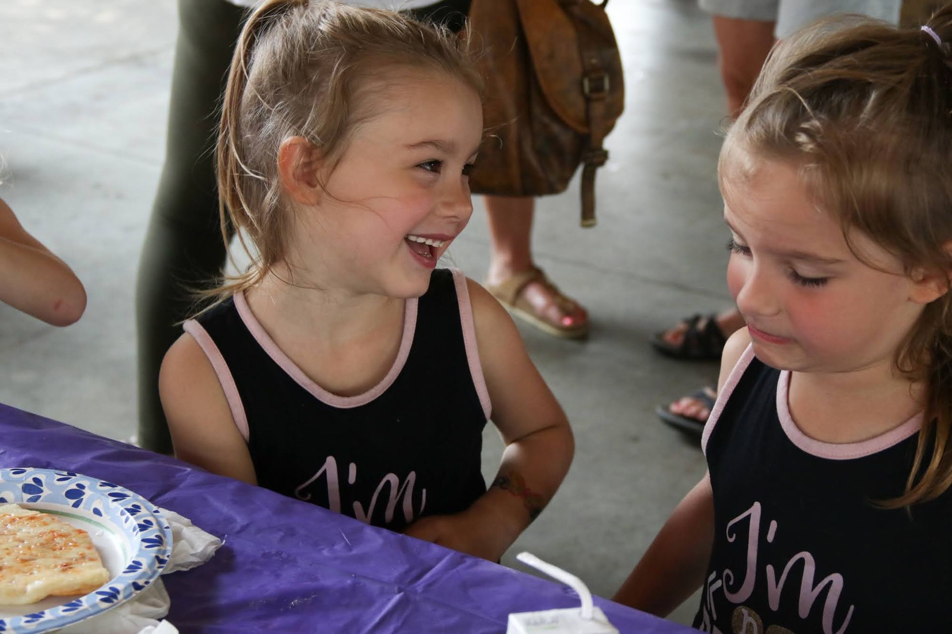 Two young girls in matching black tank tops at a table, one laughing and looking at the other.
